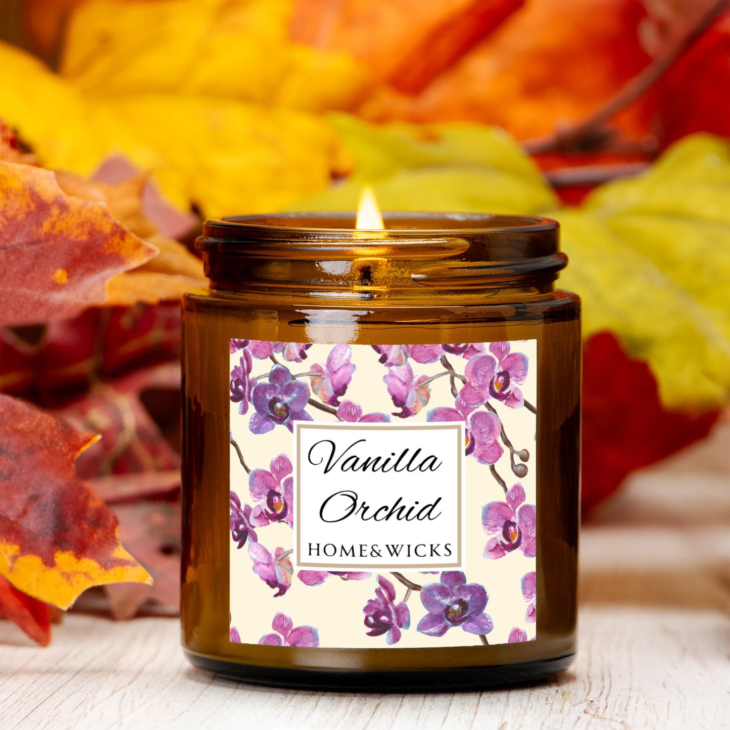 Vanilla Orchid candle in a 4 oz. amber glass candle jar with gold lid with a pink and purple orchid floral label on a white background by Home & Wicks with autumn leaves in the background