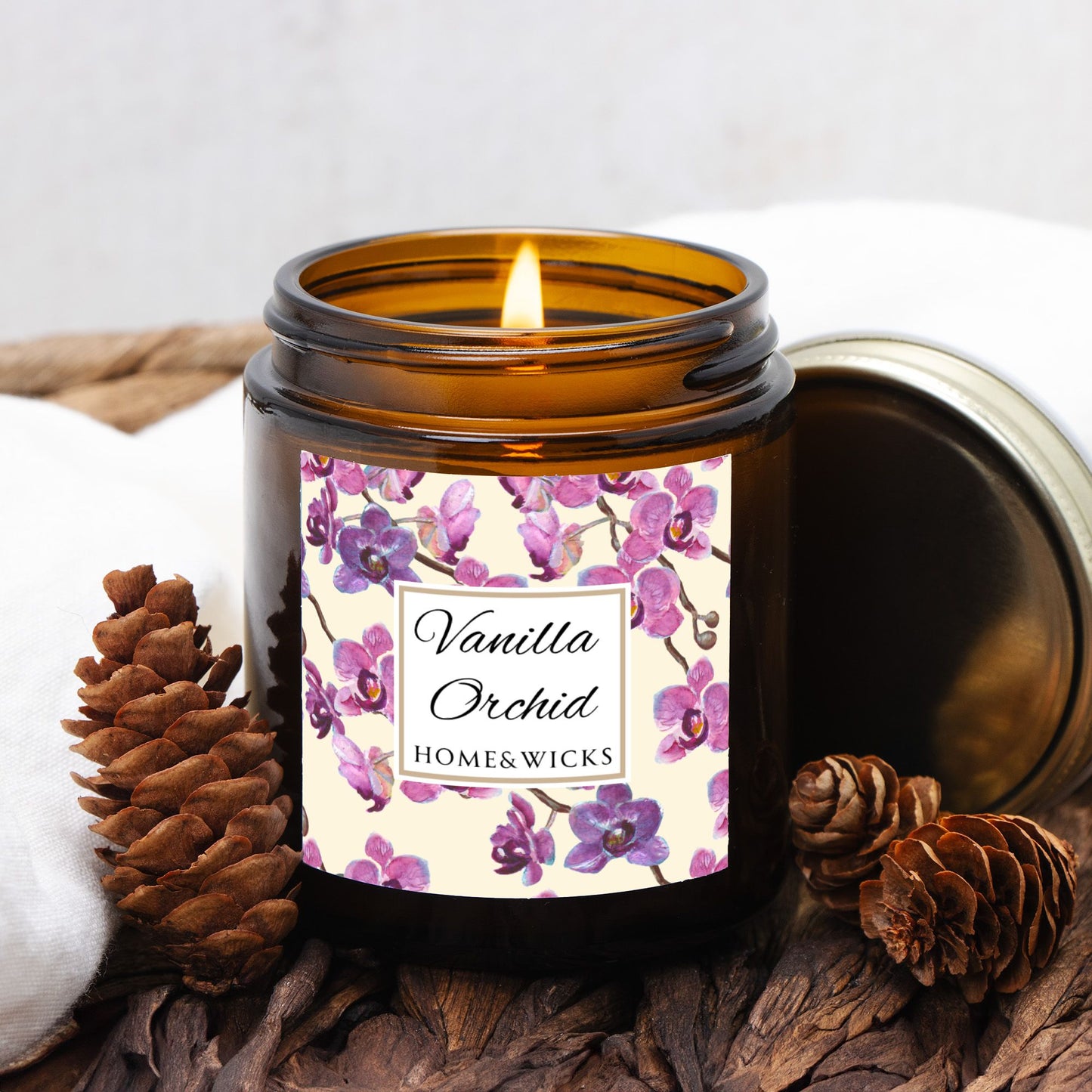 Vanilla Orchid candle in a 4 oz. amber glass candle jar with gold lid with a pink and purple orchid floral label on a white background by Home & Wicks on a wooden surface with pine cones.