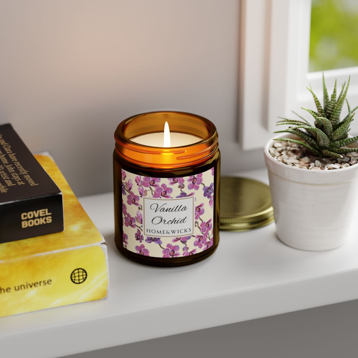 Vanilla Orchid candle in an amber jar with gold lid with a pink and purple orchid design label by Home & Wicks on a shelf with books and a plant