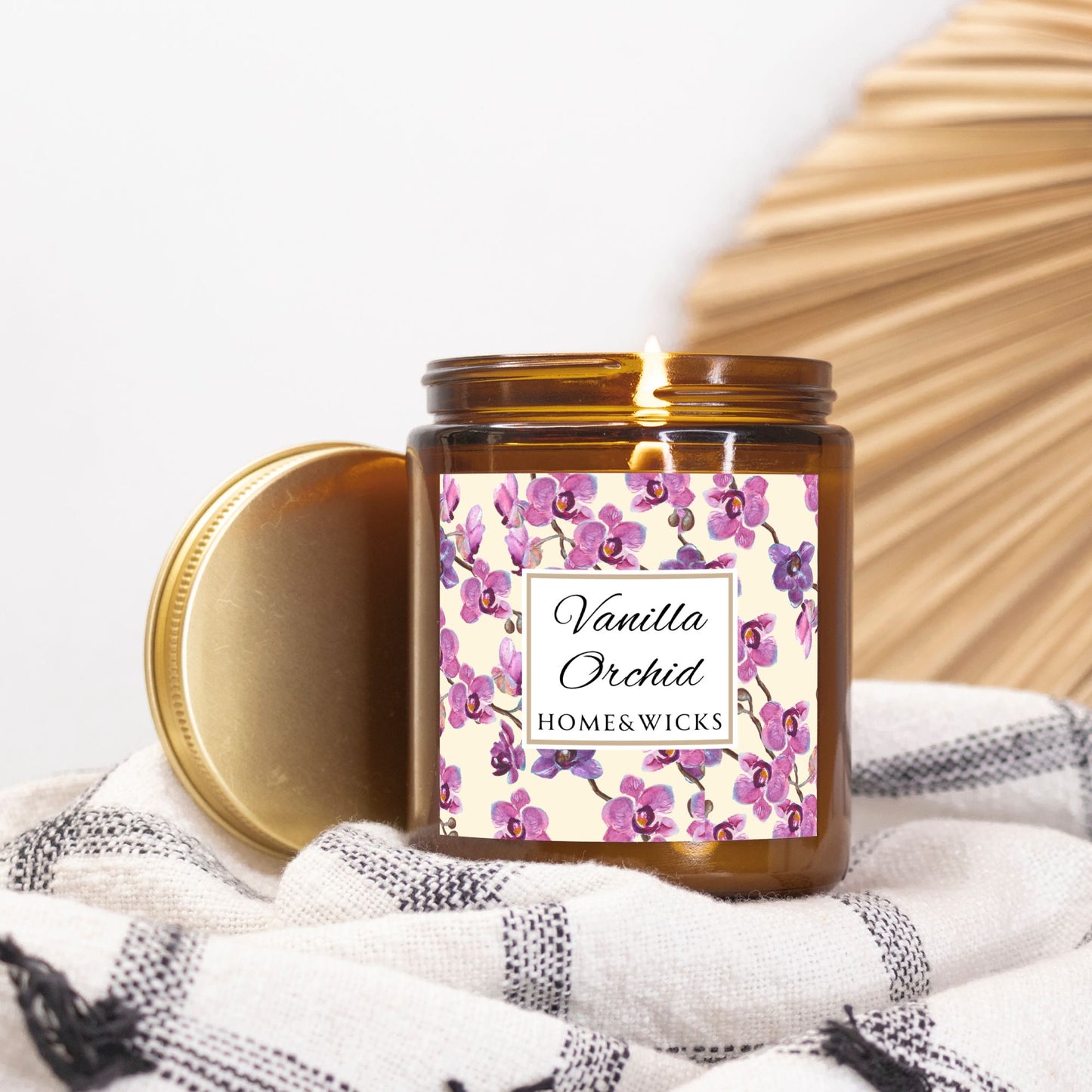 Vanilla Orchid candle in an amber jar with gold lid with a pink and purple orchid design label by Home & Wicks on a white and black checkered cloth.