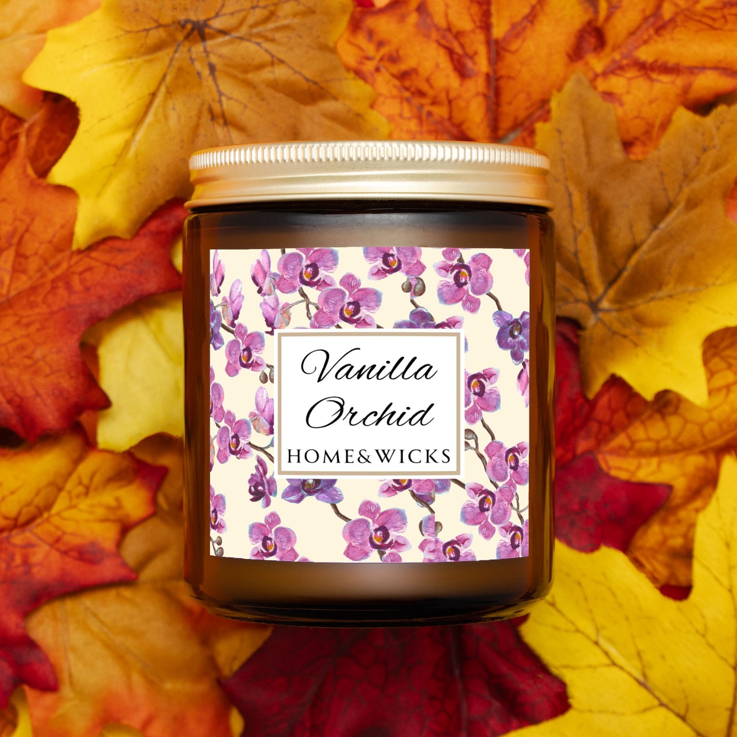 Vanilla Orchid candle in an amber jar with gold lid with a pink and purple orchid design label by Home & Wicks surrounded by fall leaves
