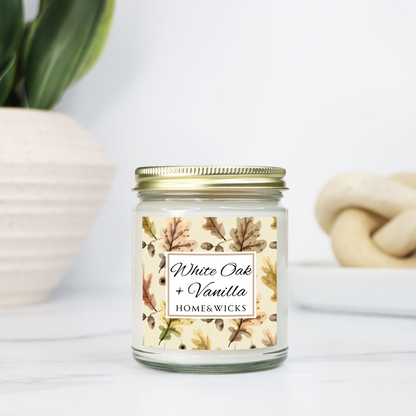 White Oak Vanilla candle in a clear jar with a gold lid labeled 'White Oak + Vanilla' with oak leaves by Home & Wicks on a white background on a light background