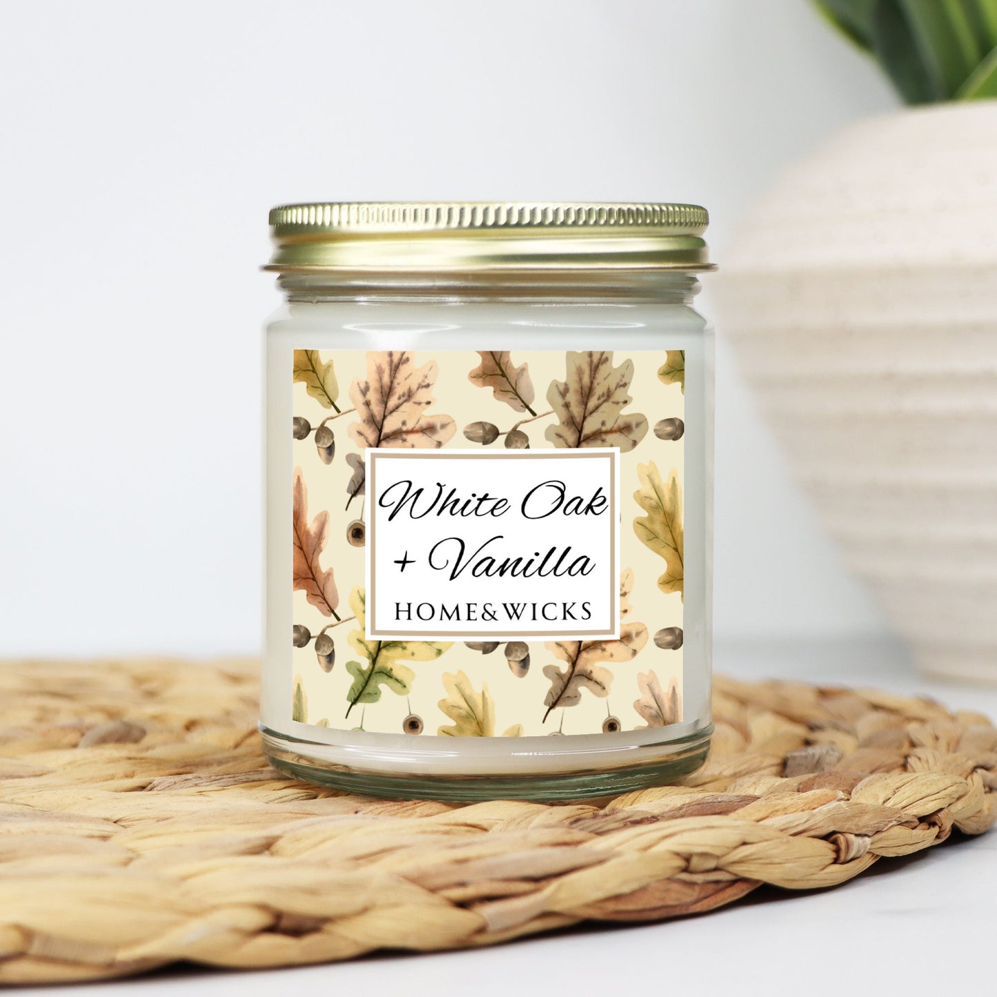 White Oak Vanilla candle in a clear jar with a gold lid labeled 'White Oak + Vanilla' with oak leaves by Home & Wicks on a white background on a woven mat with a blurred background