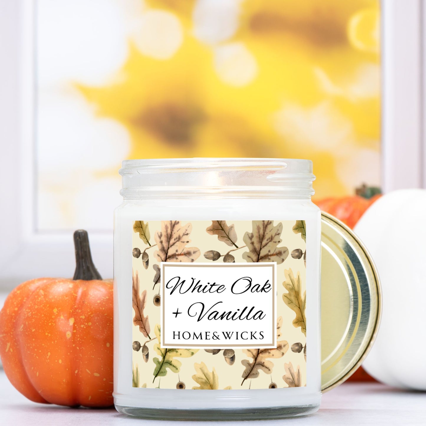 White Oak Vanilla candle in a clear jar with a gold lid labeled 'White Oak + Vanilla' with oak leaves by Home & Wicks on a white background with pumpkins in the background