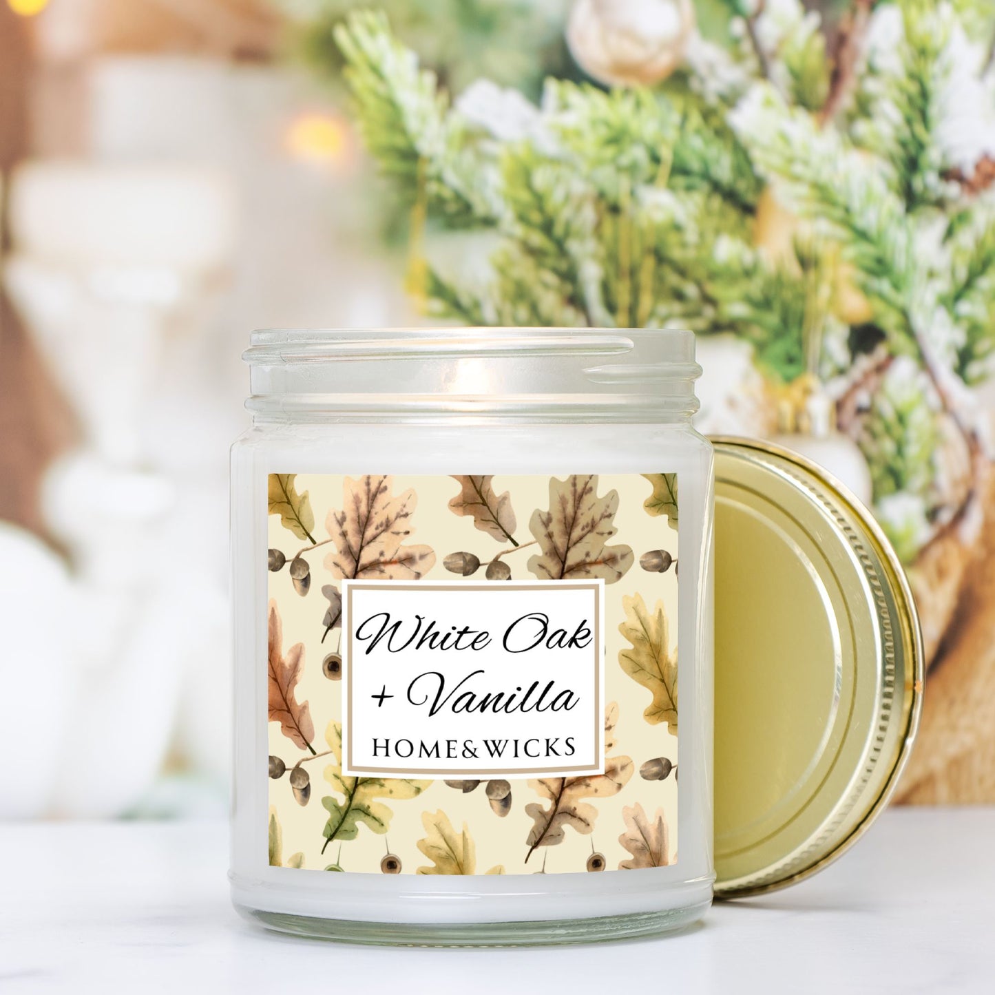 White Oak Vanilla candle in a clear jar with a gold lid labeled 'White Oak + Vanilla' with oak leaves by Home & Wicks on a white background, surrounded by greenery.