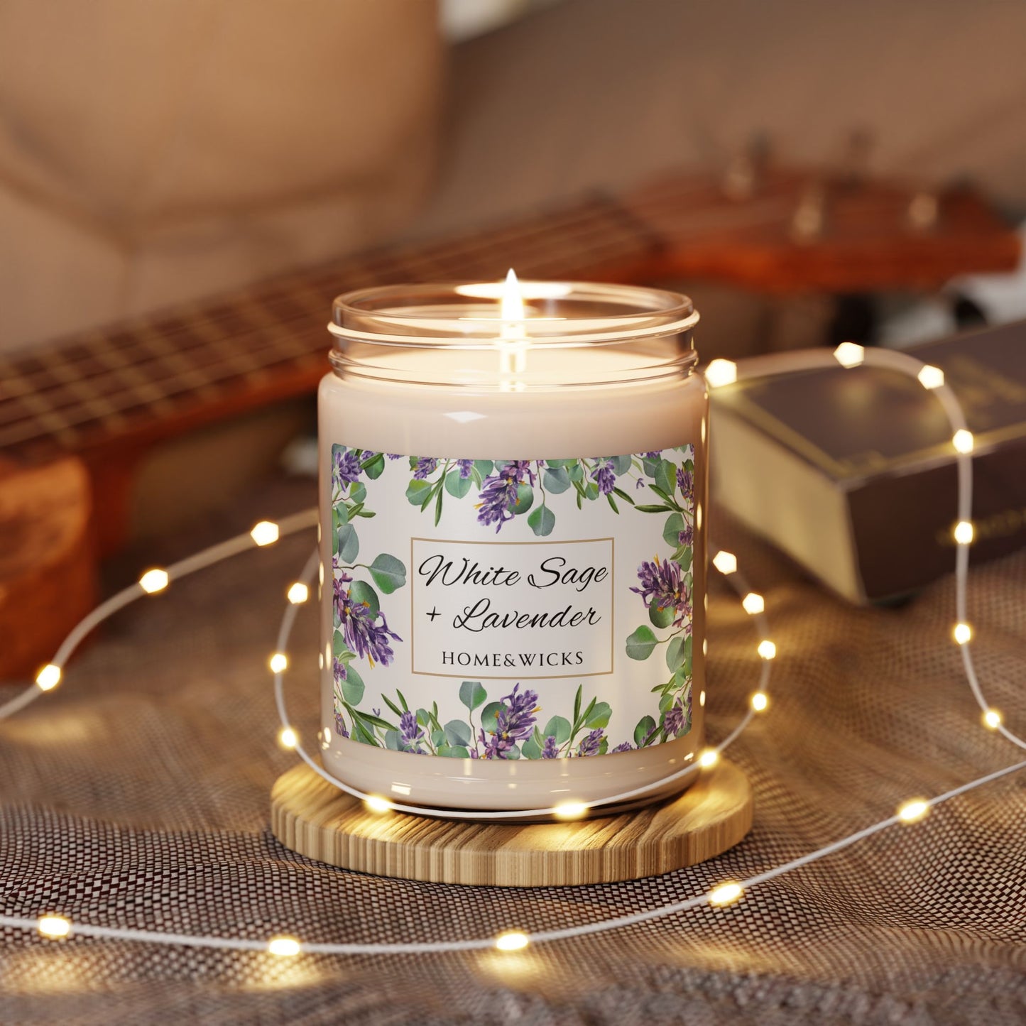 Lit candle with a floral white sage lavender label on a wooden coaster, surrounded by string lights and a guitar in the background.