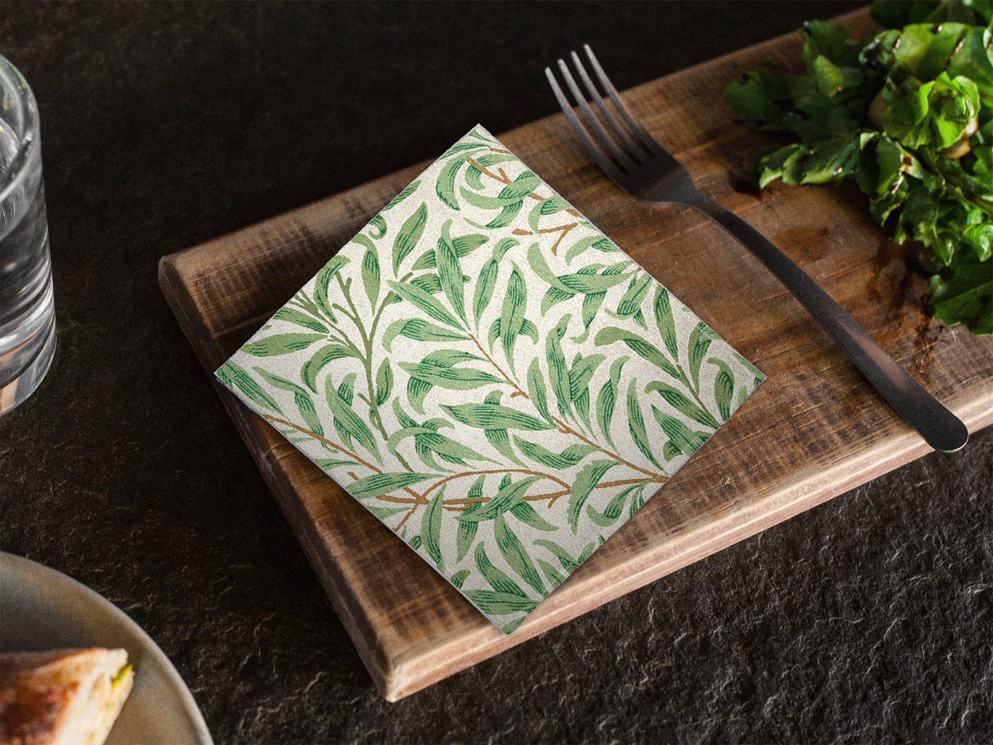 Willow Boughs 10 x 10 cocktail napkin with green leaf willow branch pattern on a wooden cutting board with a fork and water bottle in the background.
