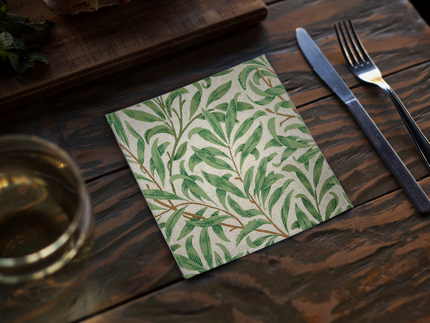 Willow Boughs 10 x 10 cocktail napkin with green leaf willow branch pattern on a wooden table with cutlery and glass.