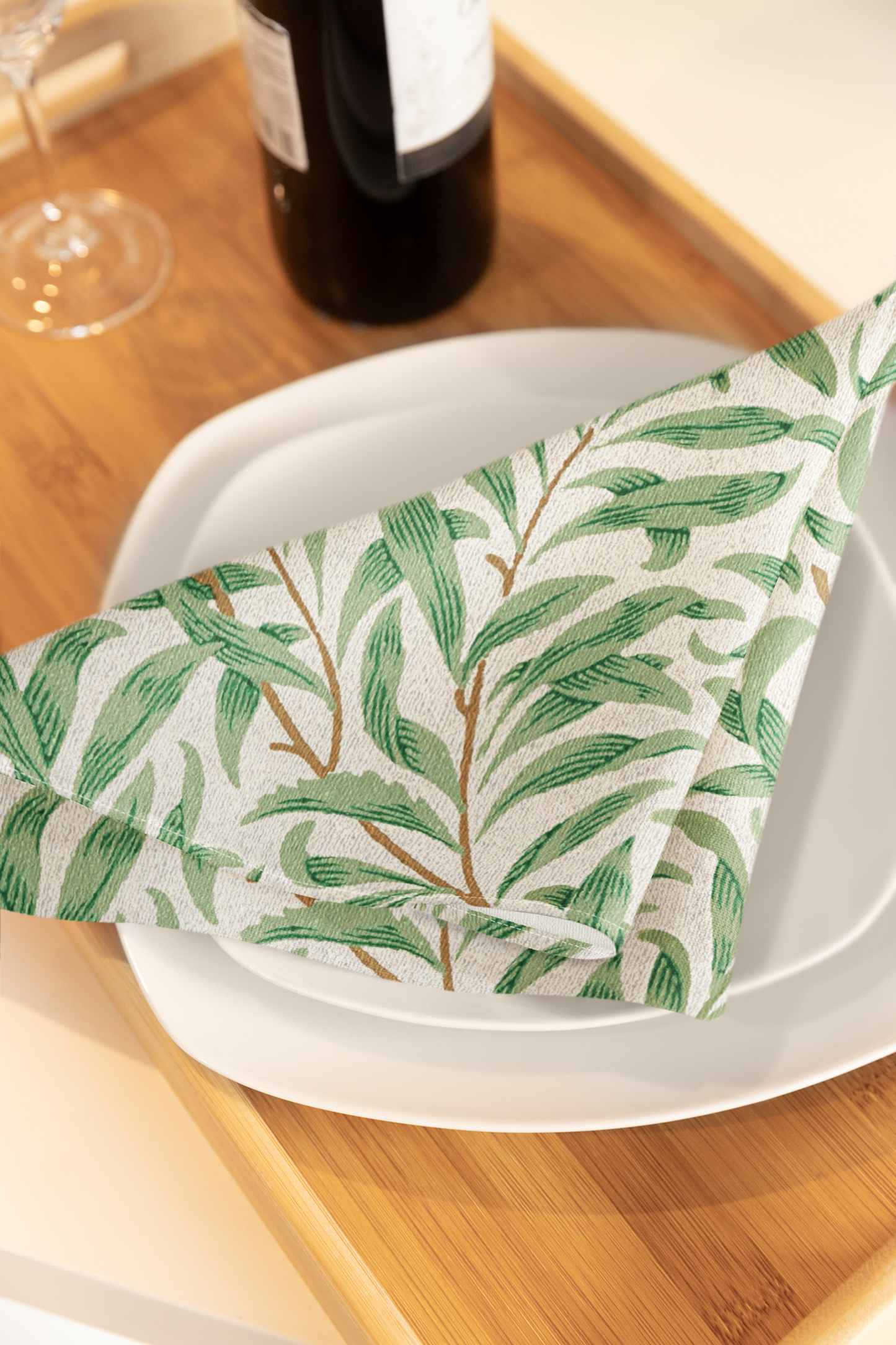 Willow Boughs 22 x 22 Napkin with green leaf willow branch pattern on a white plate with a bottle and glass in the background.