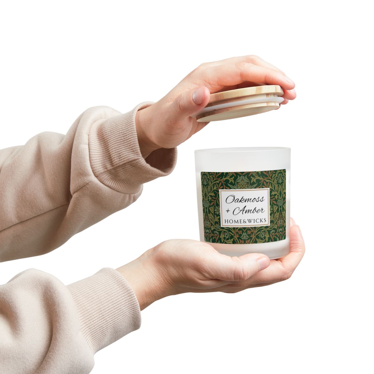 Person holding a candle Wood wick Oakmoss & Amber candle in a frosted glass jar with a green decorative label featuring the illustration by William Morris "Flower Garden" on a white background by Home & Wicks