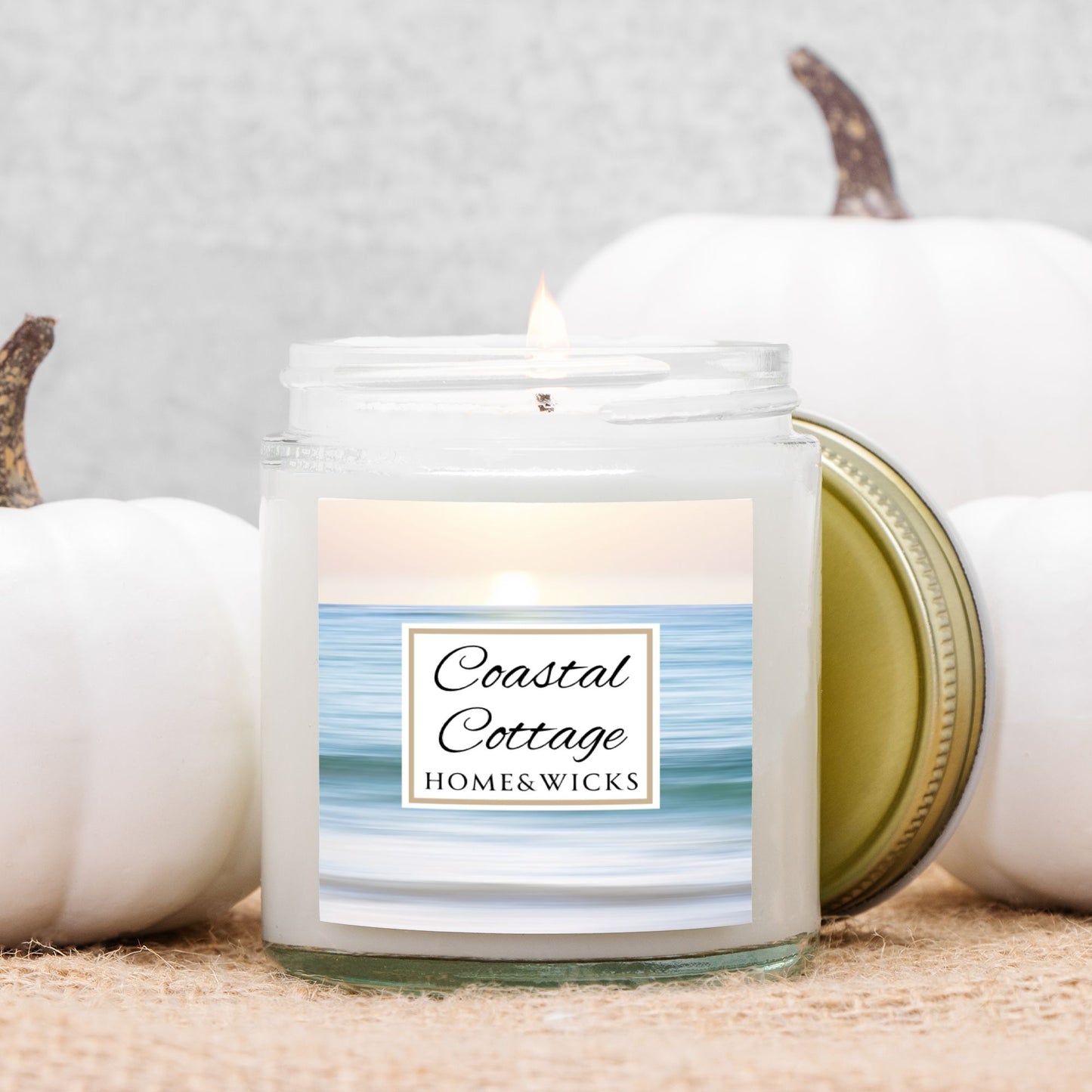 Coastal Cottage candle jar with a label reading 'Coastal Cottage' by 'Homes & Wicks' with a calm ocean shore image on a white background surrounded by white pumpkins on a neutral background.