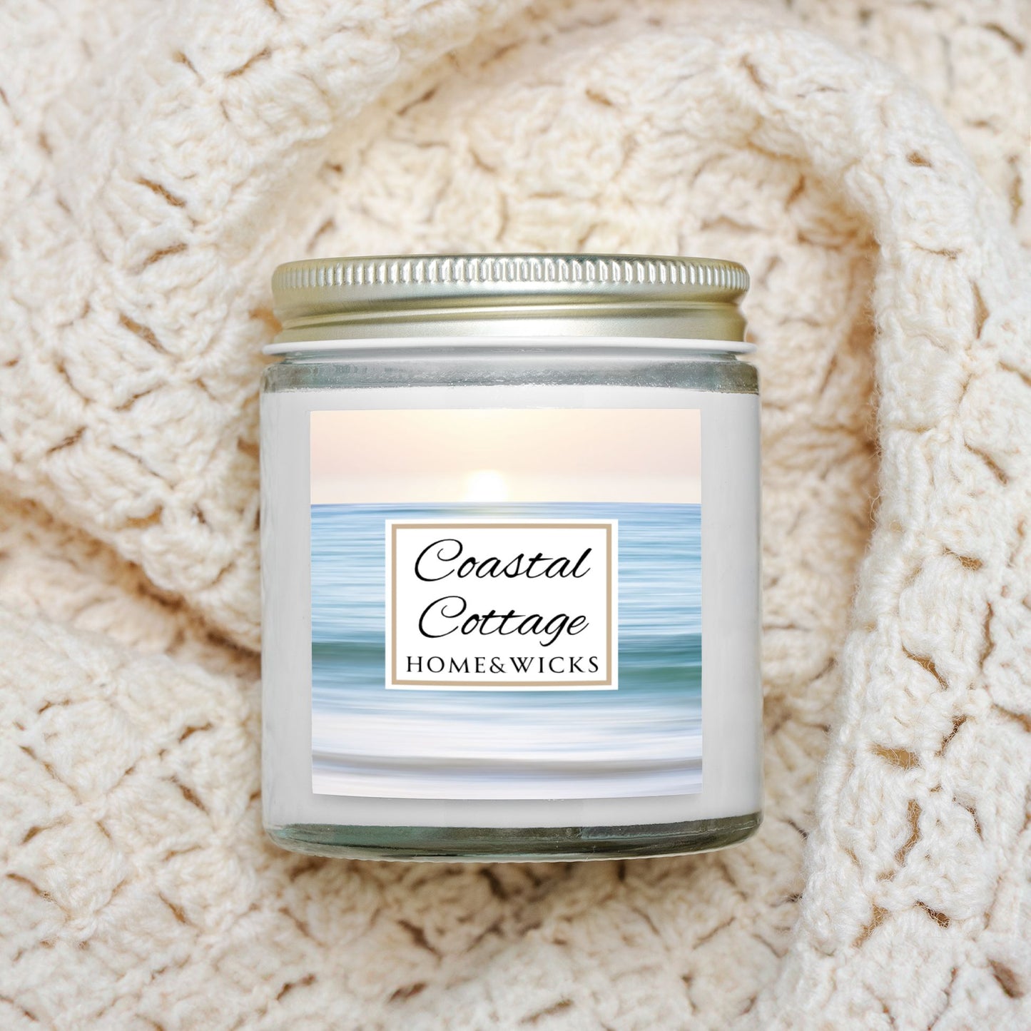 Coastal Cottage candle jar with a label reading 'Coastal Cottage' by 'Homes & Wicks' with a calm ocean shore image on a white background on a textured beige background