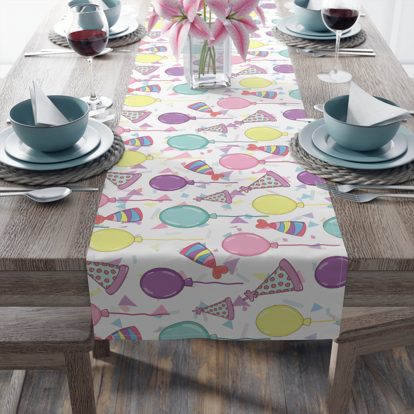 Table runner with birthday hats, confetti and purple, yellow, blue and pink balloons on a white background on a dark wood table with blue plates and bowls