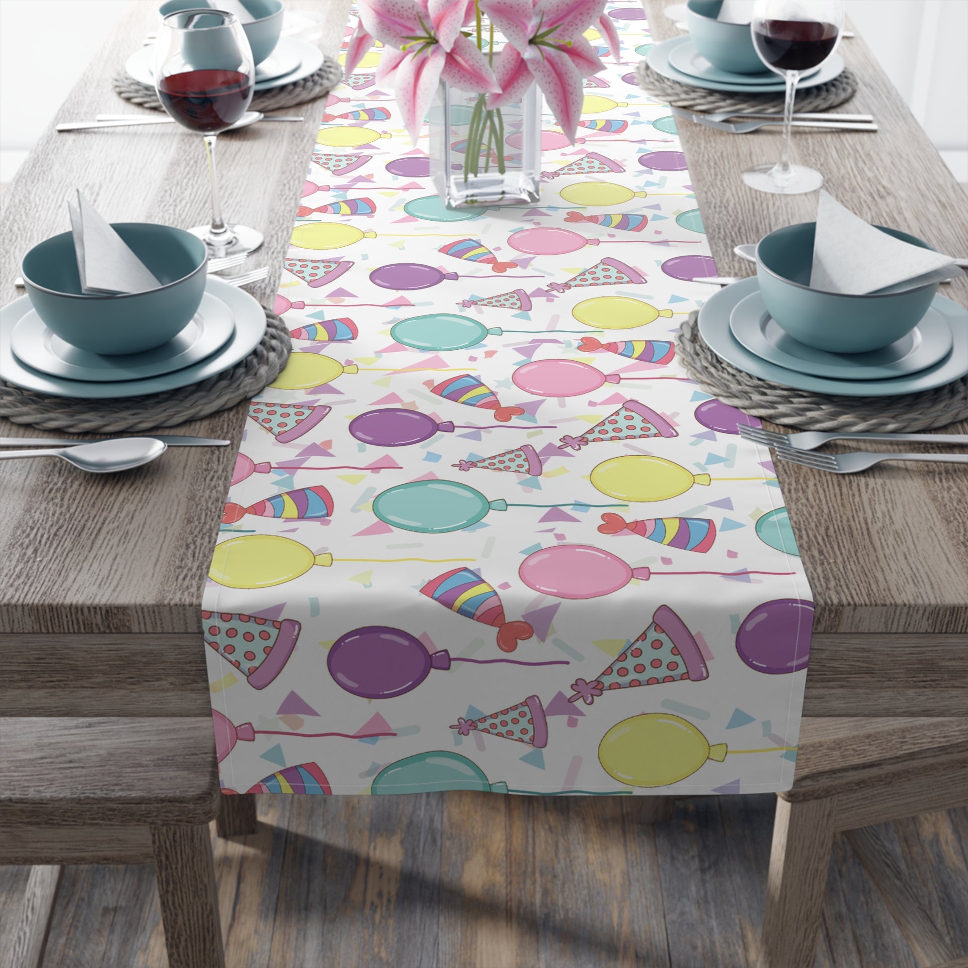 Table runner with birthday hats, confetti and purple, yellow, blue and pink balloons on a white background on a dark wood table with blue plates and bowls