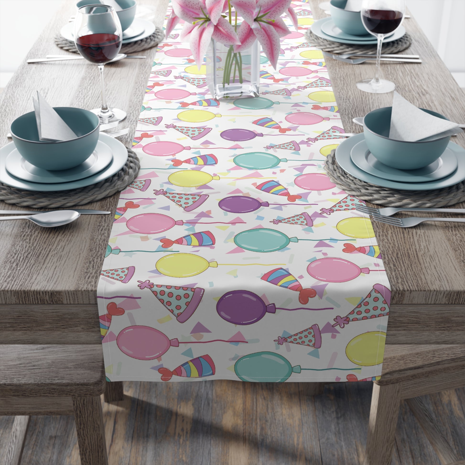 Table runner with birthday hats, confetti and purple, yellow, blue and pink balloons design on a white background on a dark wood table with blue plates and bowls 