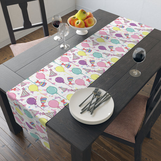 Table runner with birthday hats, confetti and purple, yellow, blue and pink balloons on a white background on a dark wood table 