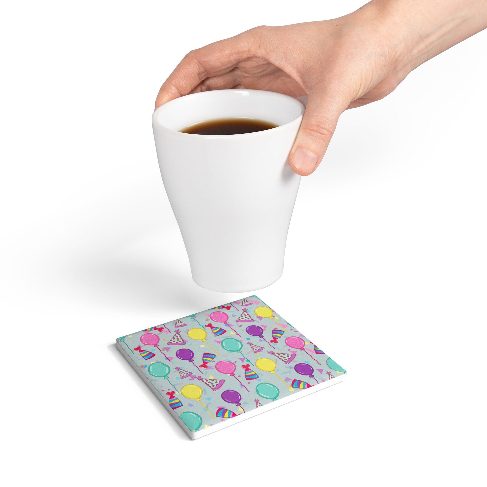 Happy Birthday square ceramic tile coaster with with birthday hats, confetti and balloons pattern with a blue-green light cyan background on white background with person placing coffee cup on coaster