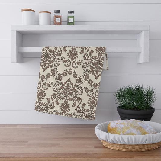 Decorative baroque mocha tea towel with brown pattern on a wooden surface with a plant and shelf in the background.