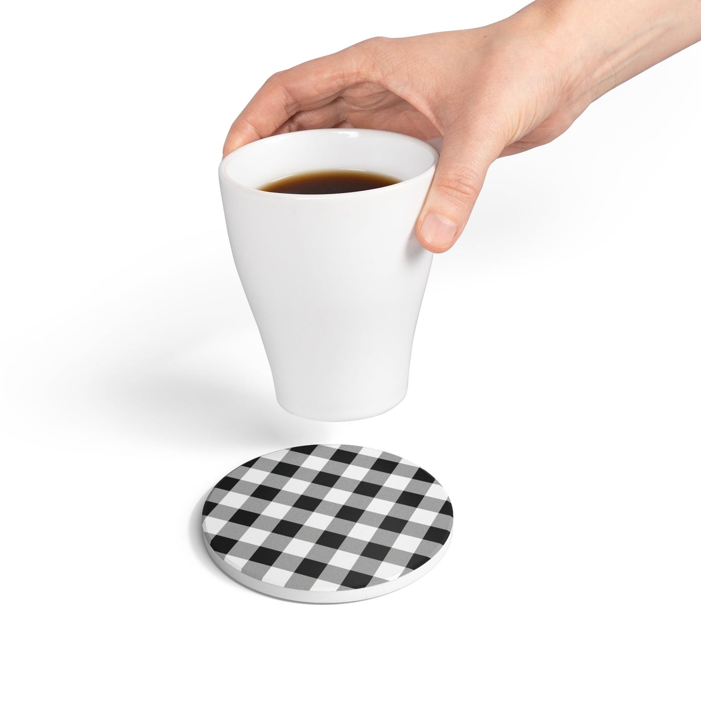 Hand holding a white mug with coffee over a round checkered black and white gingham patterned tile coaster 