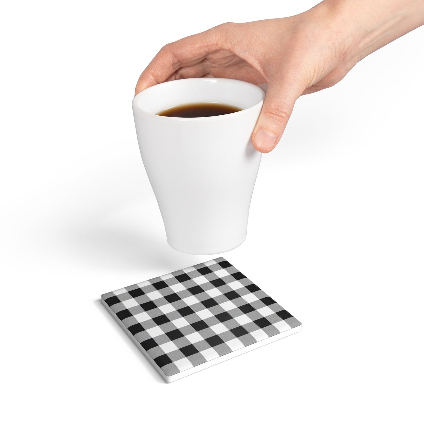 White mug with coffee held above a black and white checkered gingham square tile coaster on a white background