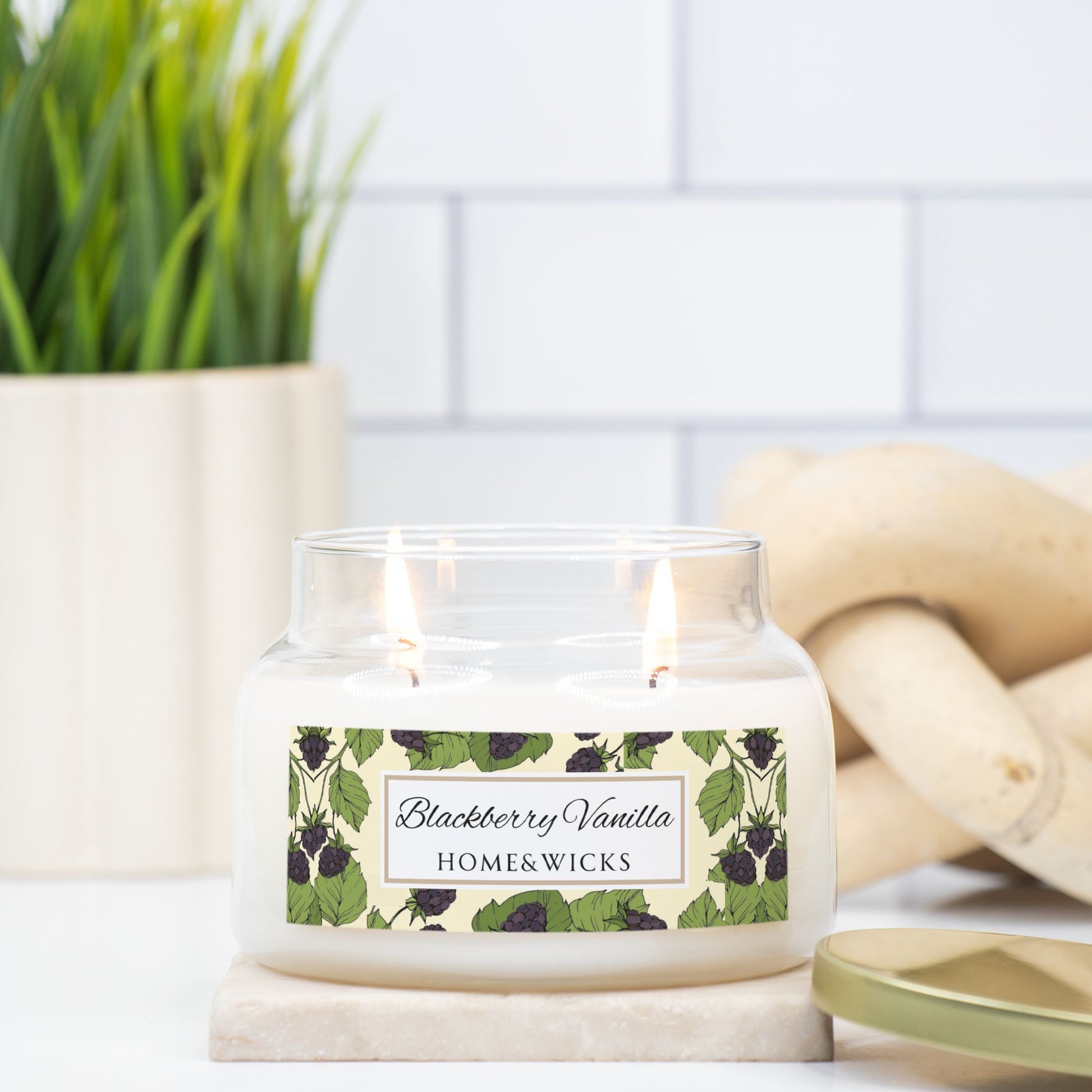 Candle with a 'Blackberry Vanilla' label on a marble surface, with a plant and wooden logs in the background.