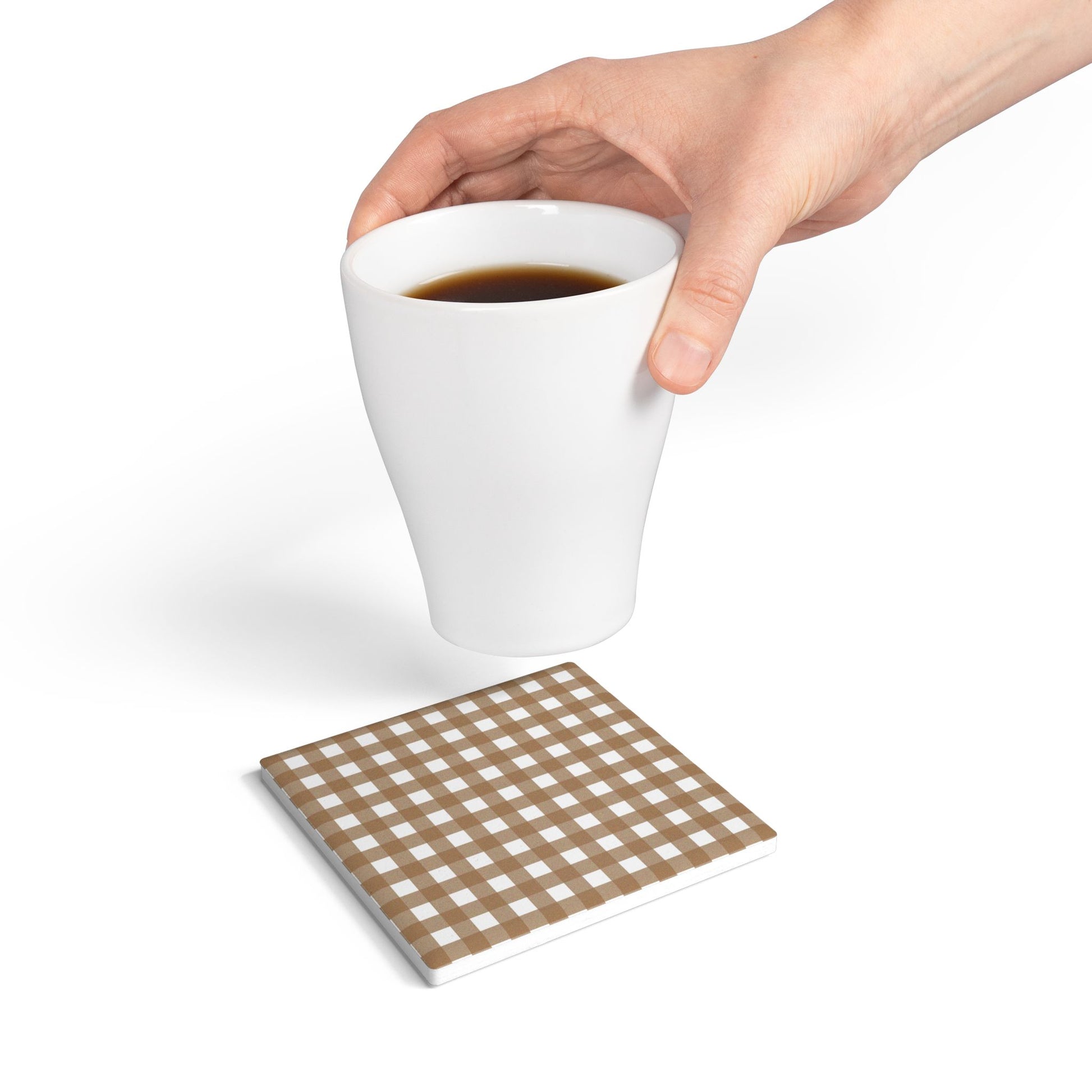 White mug with coffee held by a hand, above  a brown gingham checkered square ceramic tile coaster on a white background