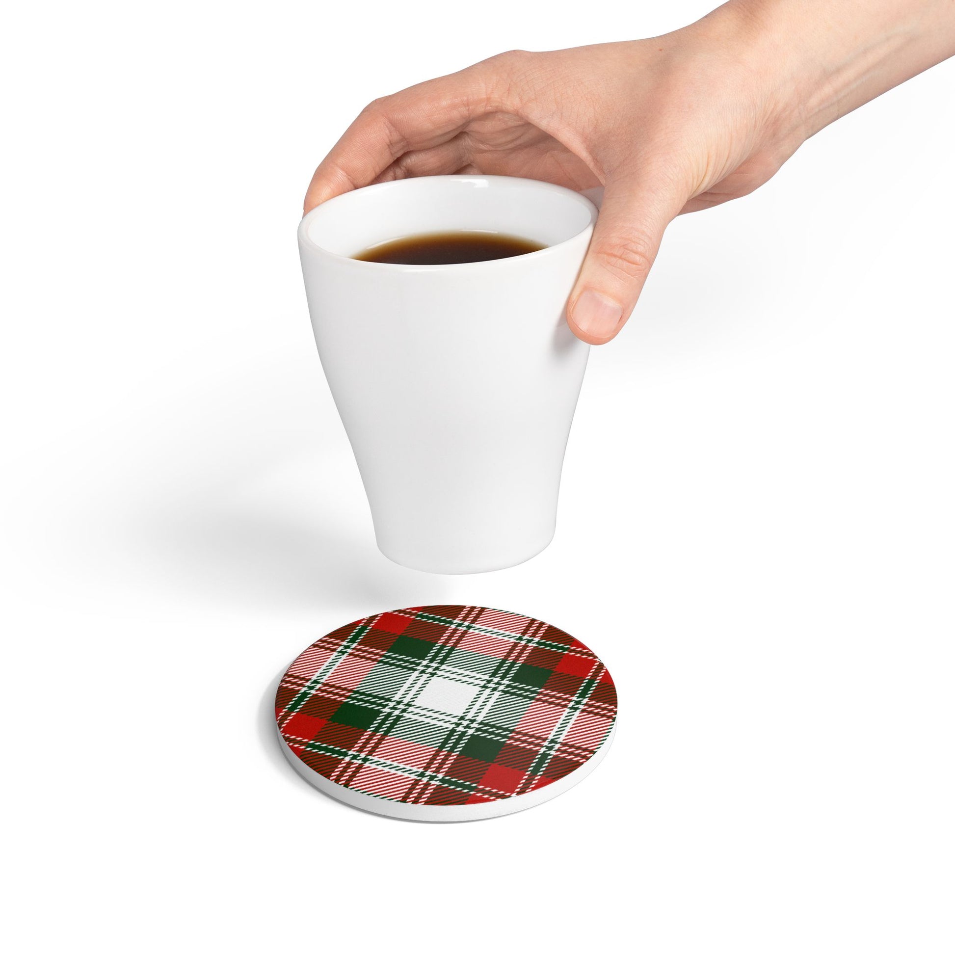 Hand holding a white mug with coffee over a green and red plaid tile coaster on a white background