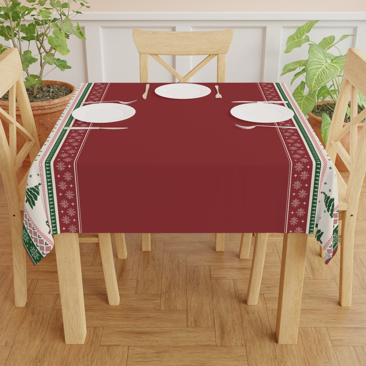 Table set with a red tablecloth with Christmas trees and snowflakes red and green pattern, white plates, and utensils on a wooden floor.
