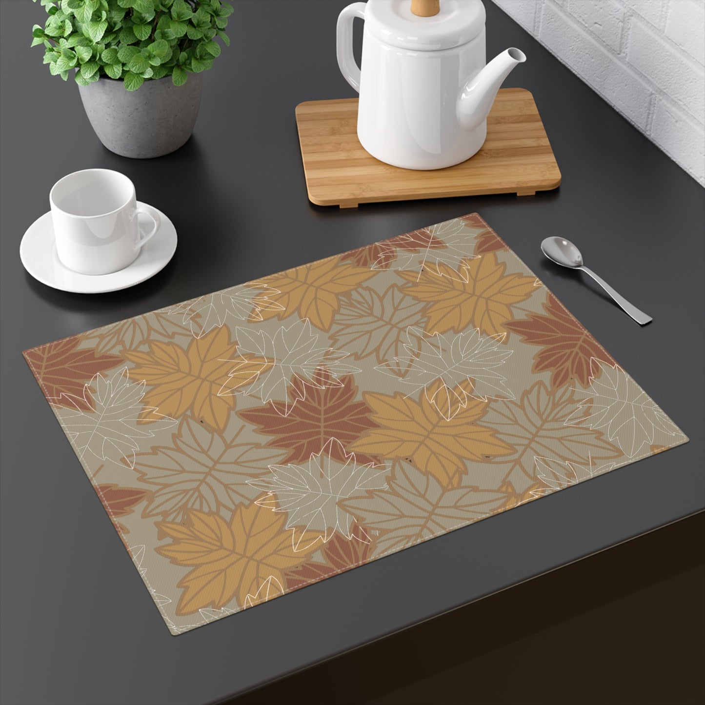 Our Earthy Leaves placemat with a brown, gold and olive green-gray autumn leaf pattern on a dark surface with tea set and plant.