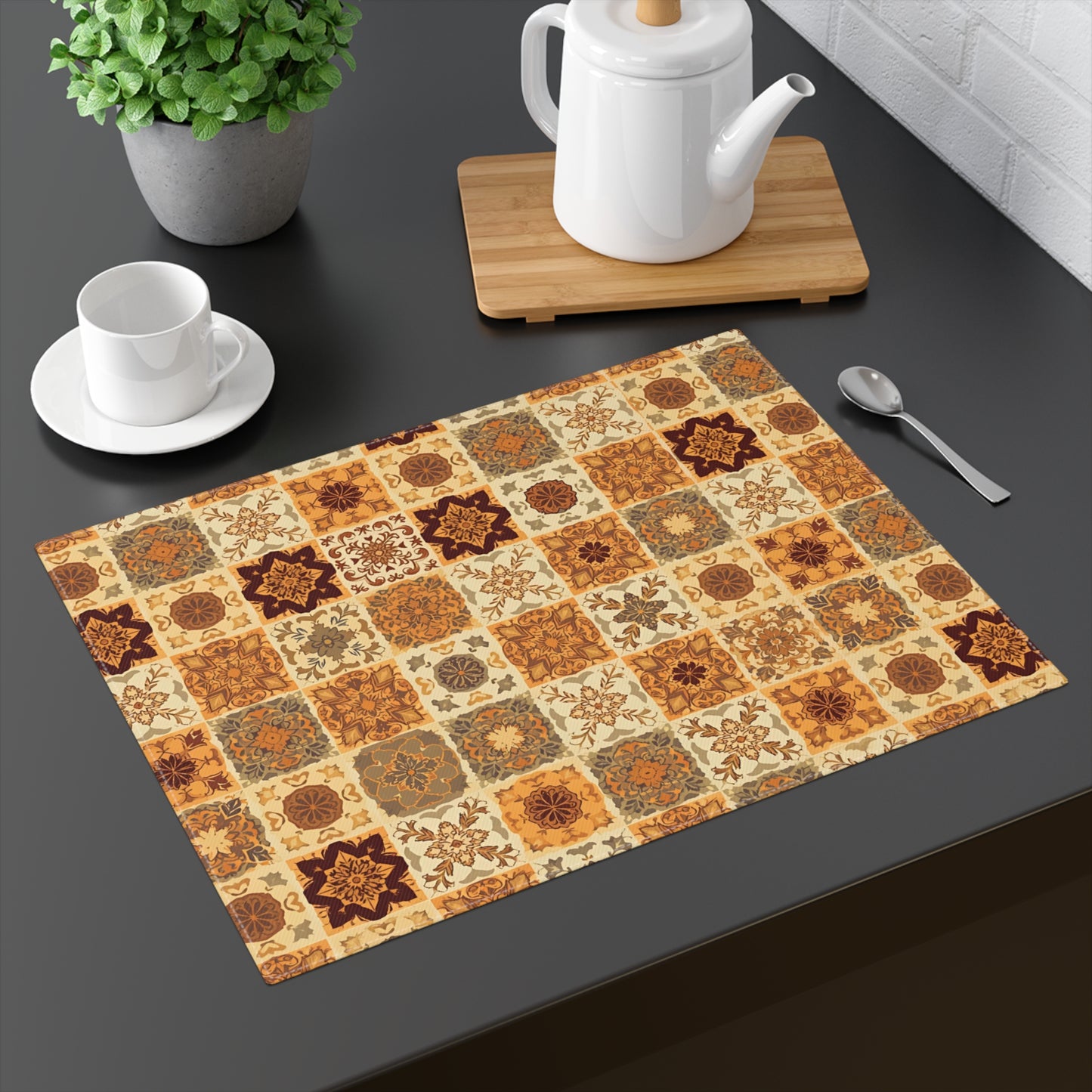 This cotton twill Fall Flowers Mosaic placemat has brown, green and gold floral square geometric pattern. This placemat is on a dark surface with a teapot, cup, and plant.