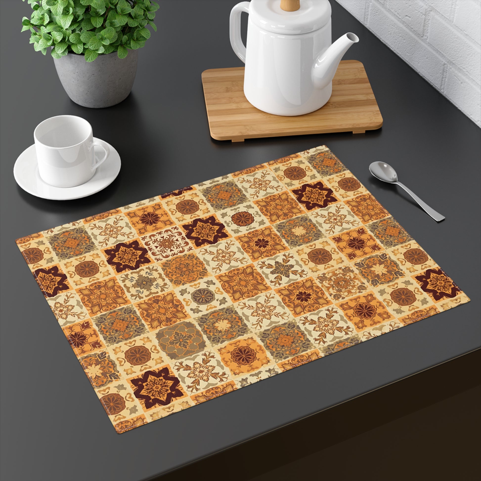 This cotton twill Fall Flowers Mosaic placemat has brown, green and gold floral square geometric pattern. This placemat is on a dark surface with a teapot, cup, and plant.