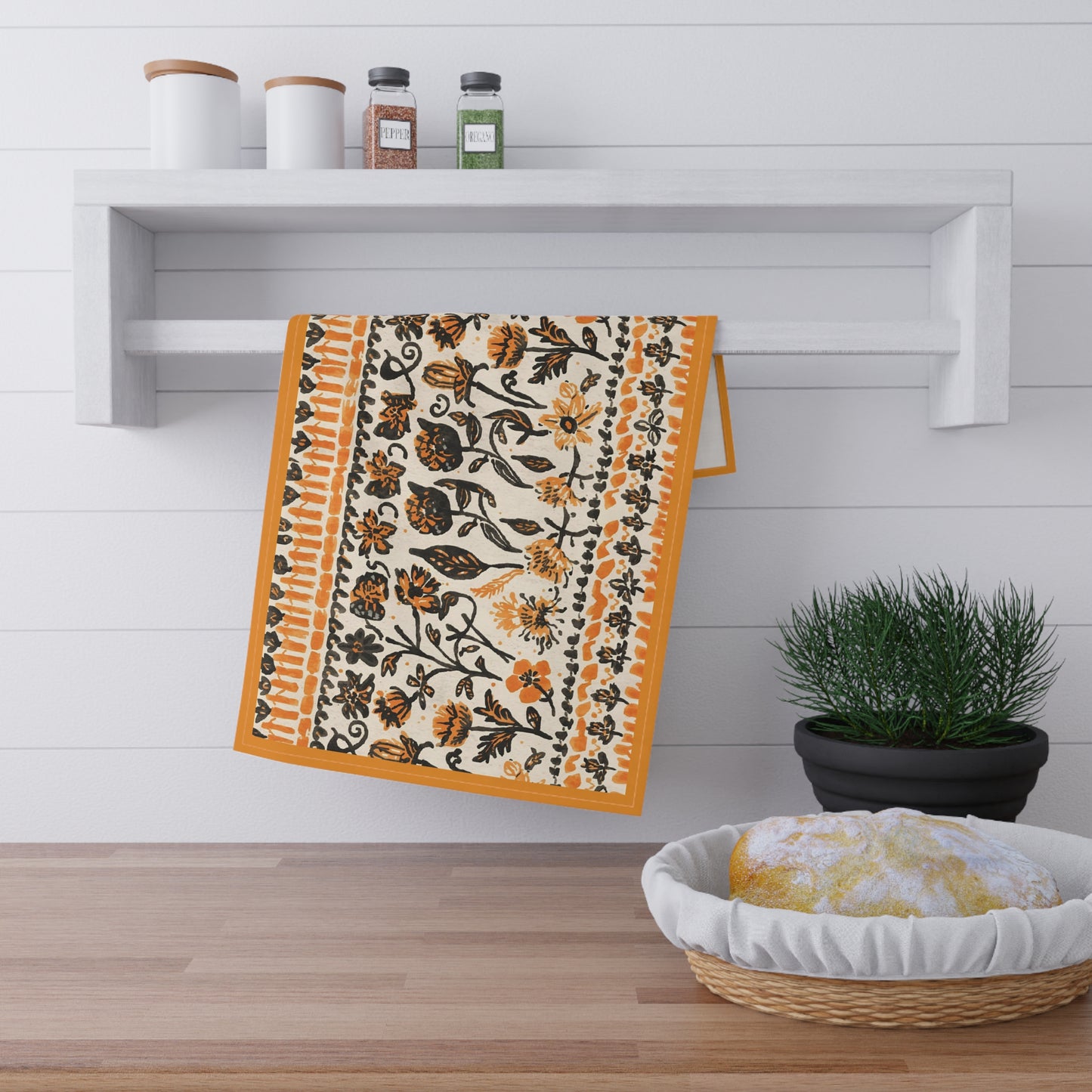 Decorative towel with black and orange floral pattern hanging on a wooden shelf with a plant and basket in the foreground.