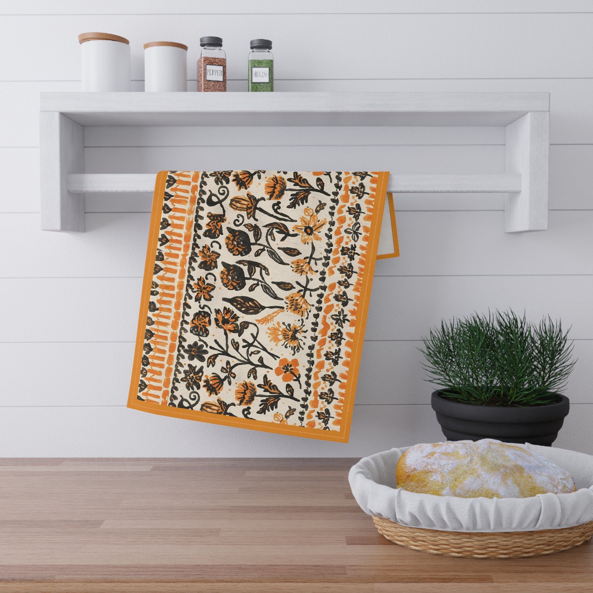 Decorative towel with black and orange floral pattern hanging on a wooden shelf with a plant and basket in the foreground.
