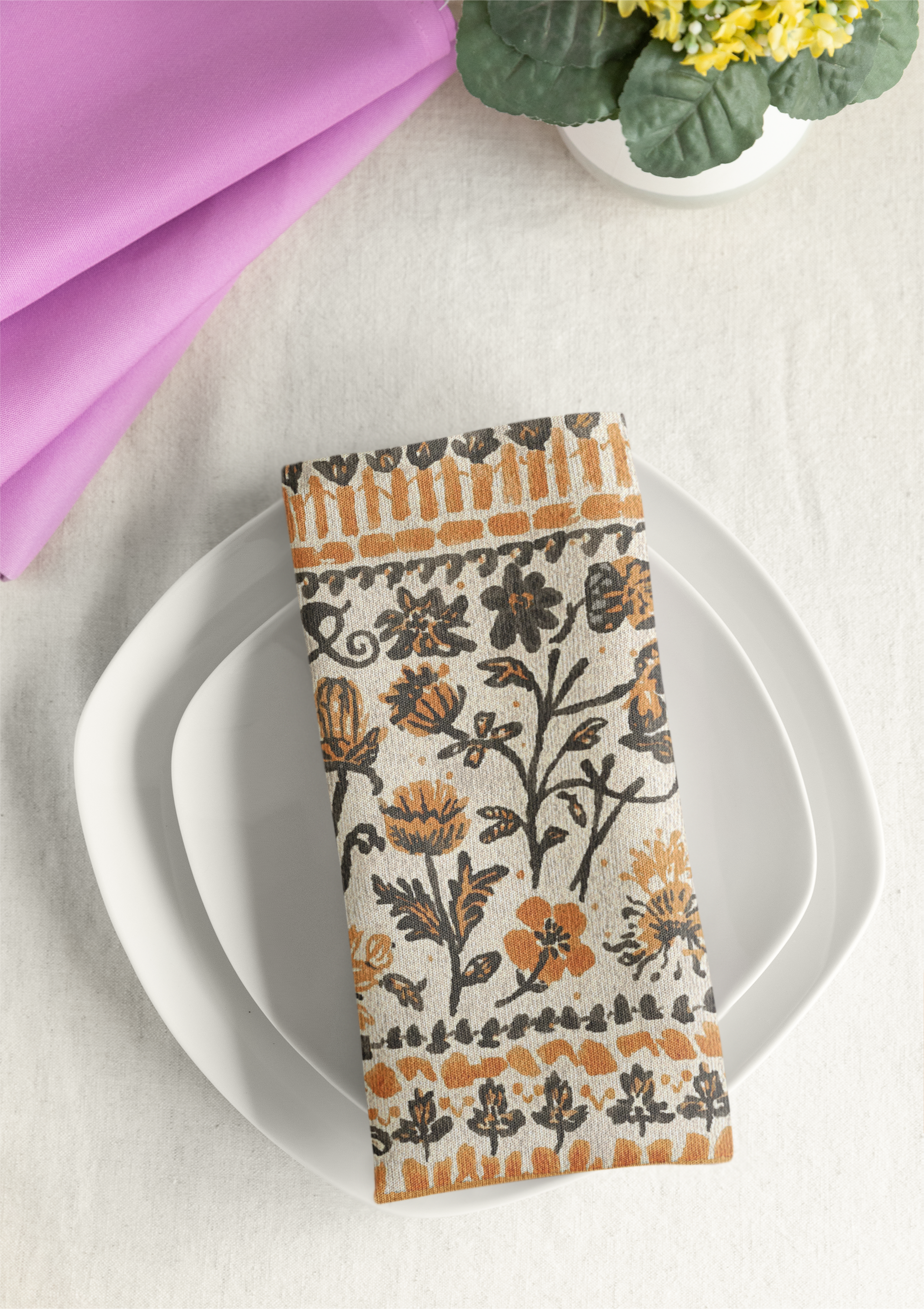 Fall Flowers napkin, with a boho orange and black floral pattern, is printed on a cotton twill reusable napkin fabric, on a white plate with a pink folded fabric and green plant in the background