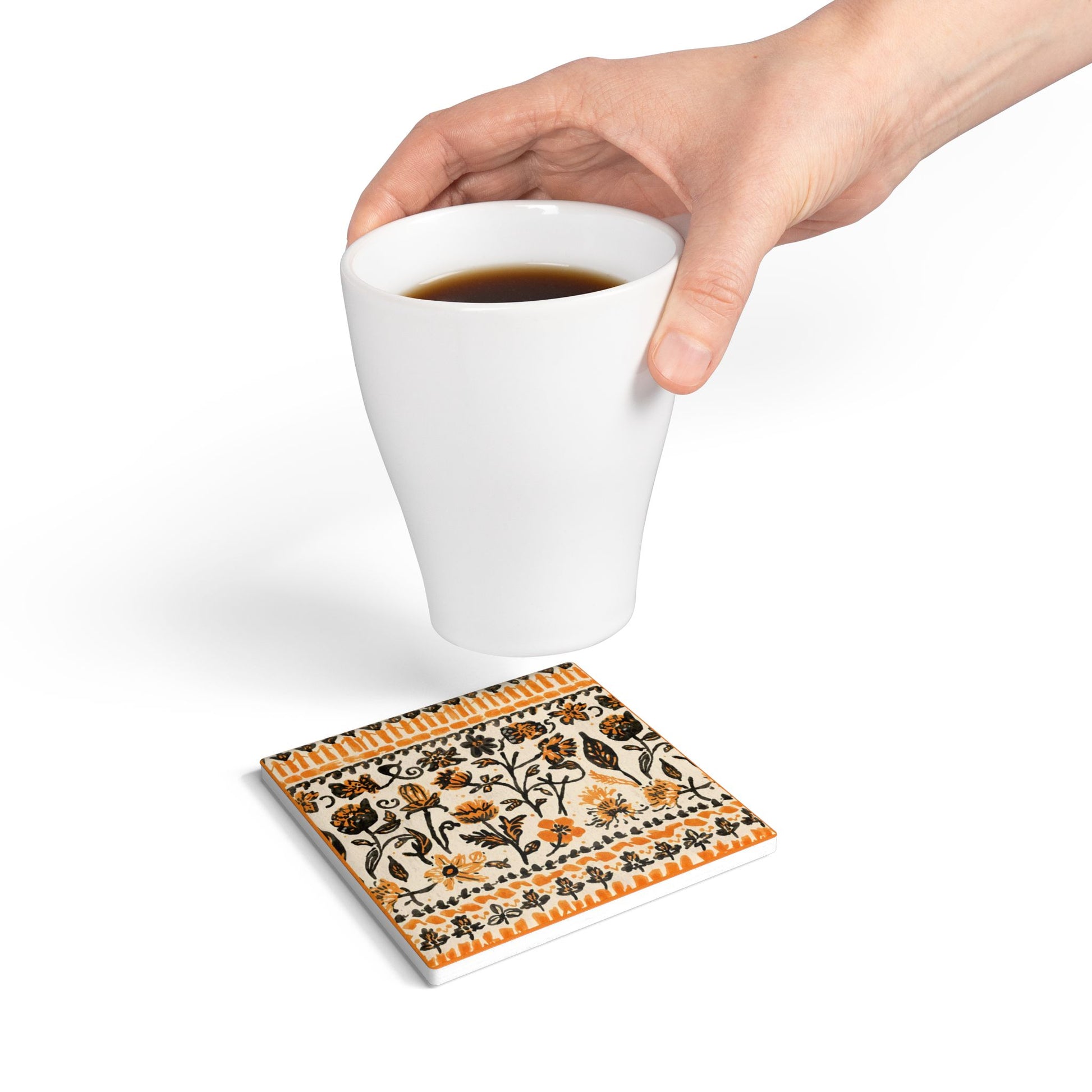 Fall Flowers ceramic tile coaster, with a boho orange and black floral pattern, is printed on the top of the ceramic tile coaster, and sits under a hand holding a white mug on a white background