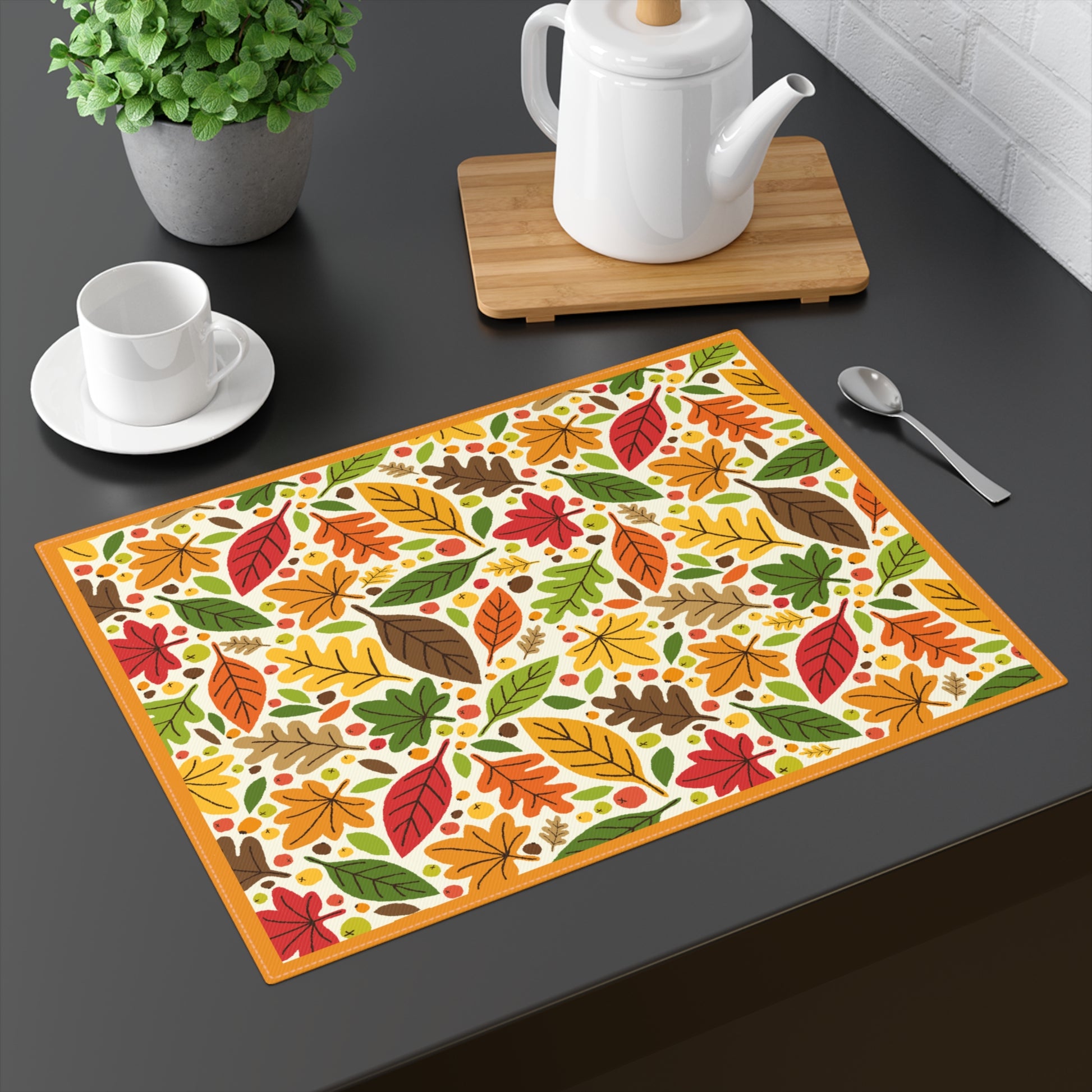 Fall Leaves placemat with autumn leaf pattern on the placemat's cotton twill fabric,  on a dark table with a teapot, cup, and spoon.
