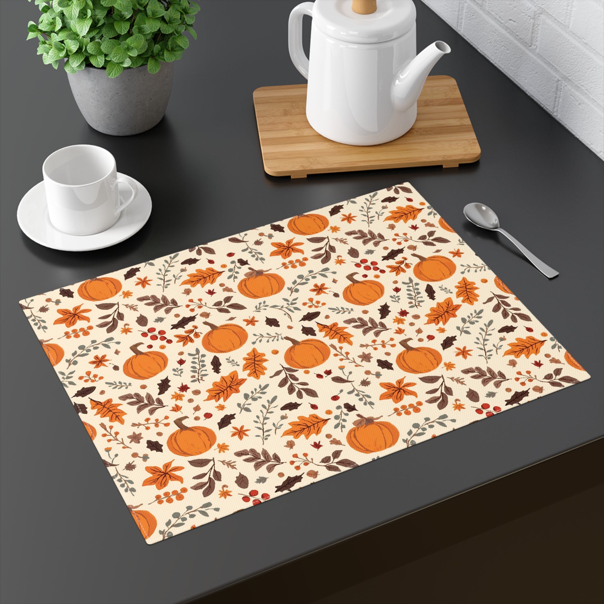 Fall Leaves and Pumpkins placemat has brown and orange fall leaves with orange pumpkins on a beige background, printed on the table runner cotton twill  fabric, shown on a dark gray table with a teapot and cup.