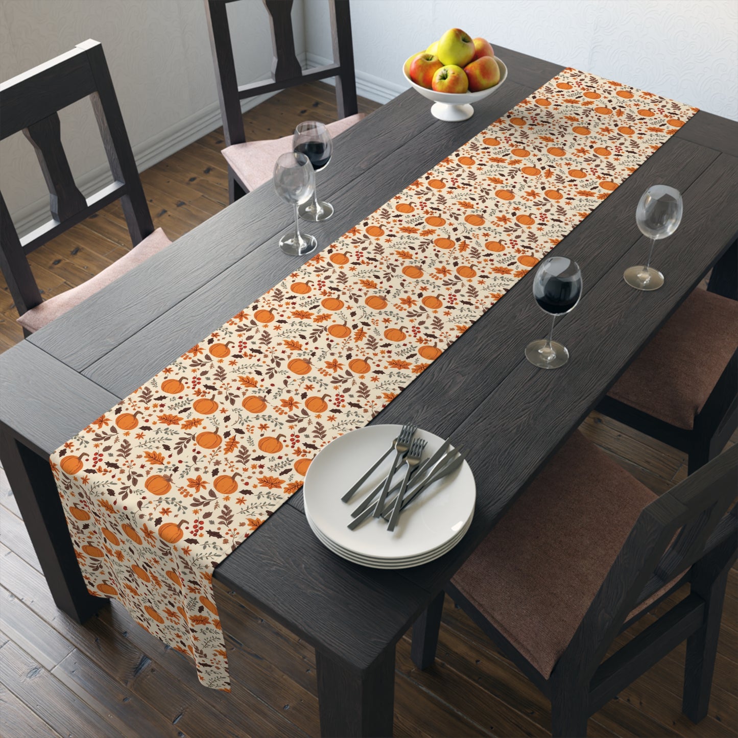 Fall Leaves and Pumpkins table runner has brown and orange fall leaves with orange pumpkins with beige background design printed on the table runner shown on a dining table with plates, and glasses in a room.