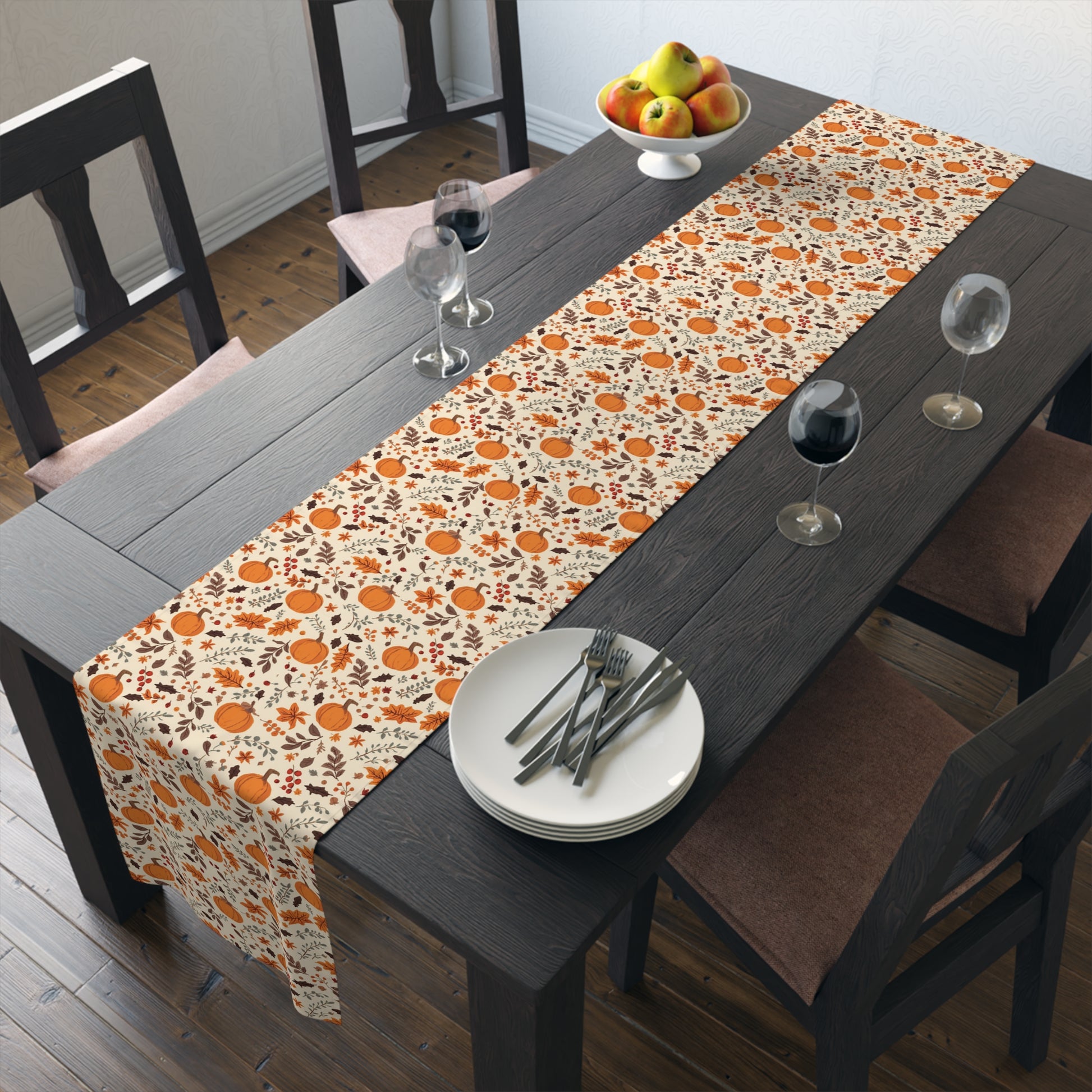 Fall Leaves and Pumpkins table runner has brown and orange fall leaves with orange pumpkins with beige background design printed on the table runner shown on a dining table with plates, and glasses in a room.