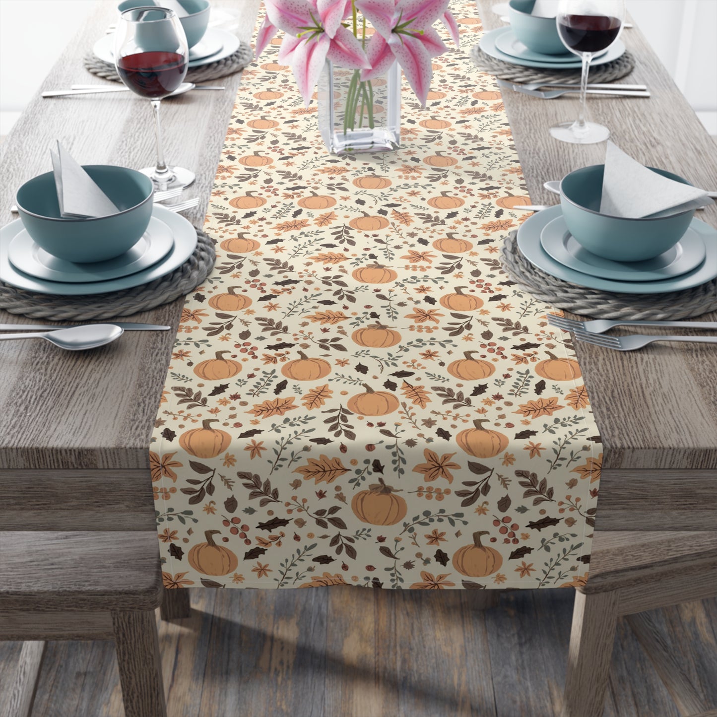 Fall Leaves and Pumpkins table runner has brown and orange fall leaves with orange pumpkins with a beige background design printed on the table runner shown on a table setting on a wooden table.