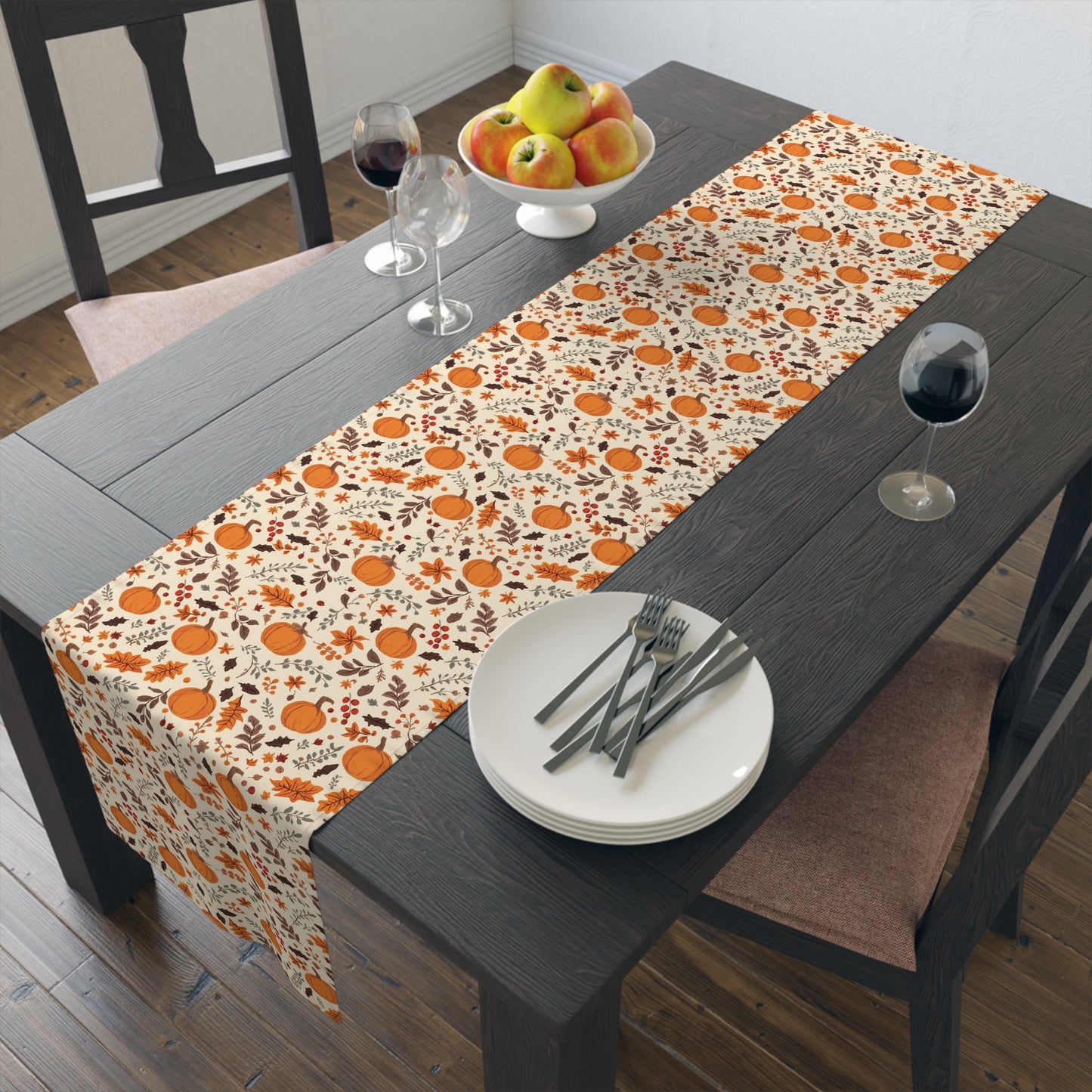 Fall Leaves and Pumpkins table runner has brown and orange fall leaves with orange pumpkins with beige background printed on the table runner shown on dining table with plates, cutlery, and glasses.