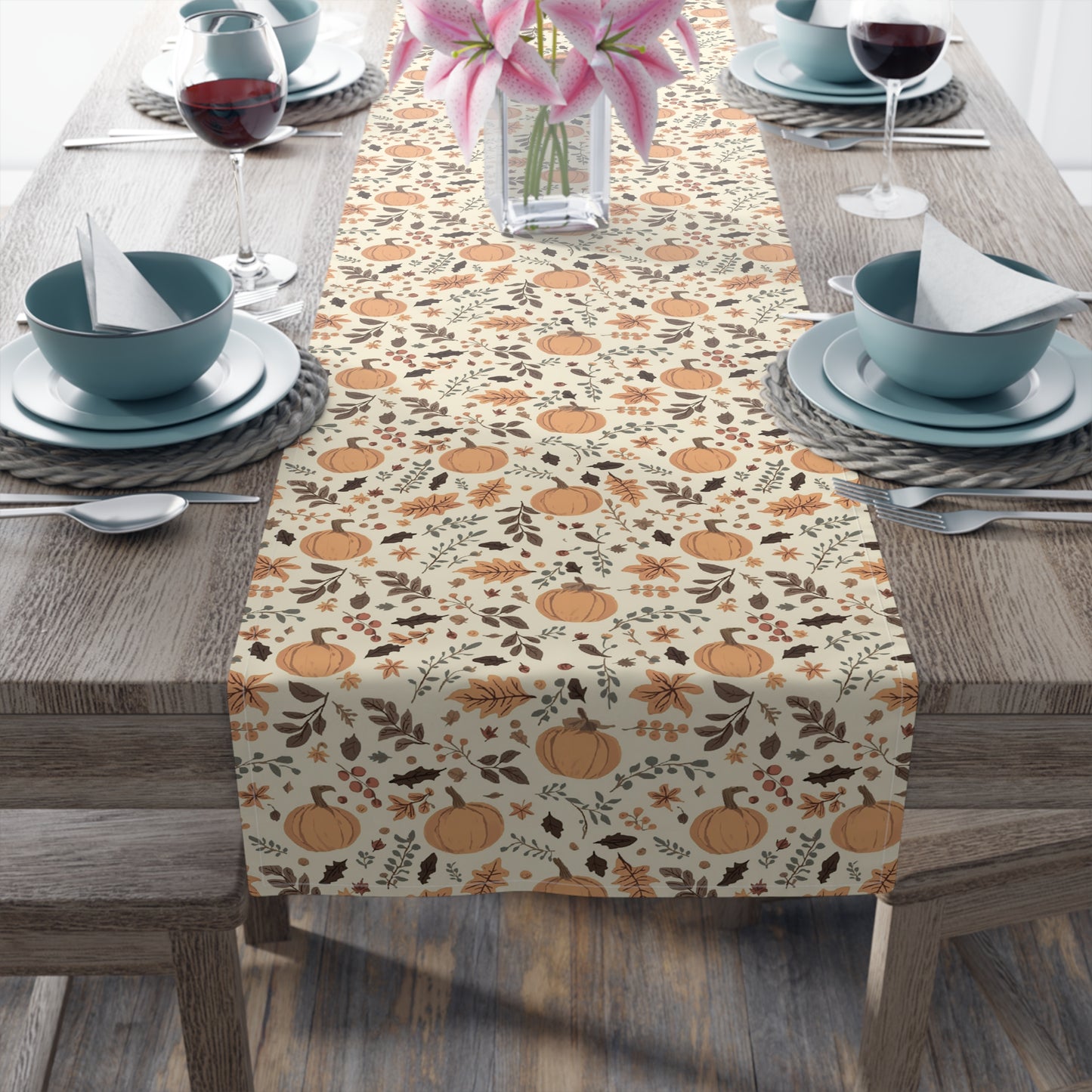 Fall Leaves and Pumpkins table runner has brown and orange fall leaves with orange pumpkins on a beige background design printed on the table runner shown on a table setting on a wooden table.