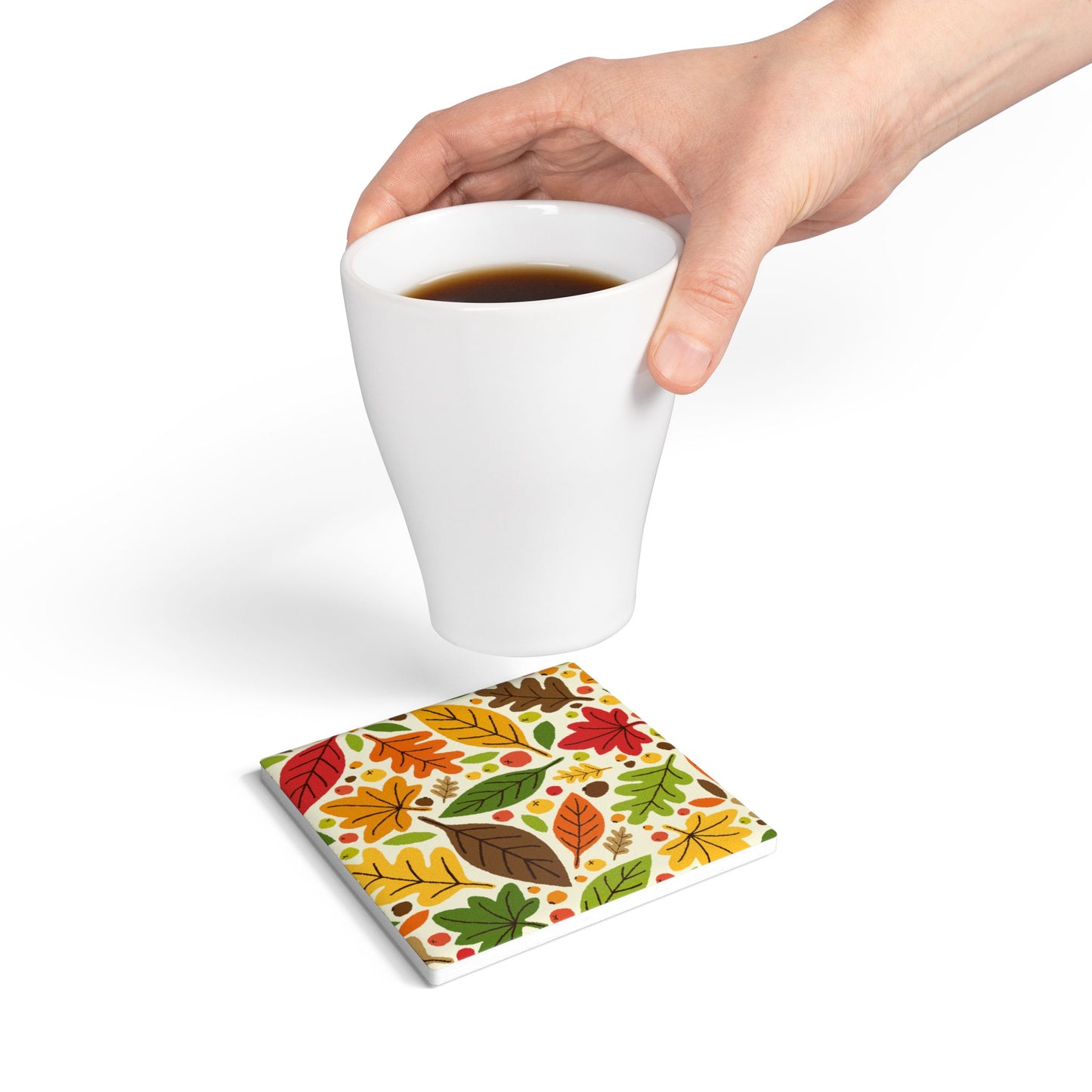 Hand holding a white mug over our square Fall Leaves ceramic tile coaster with a colorful leaf-patterned on top of coaster on a white background.