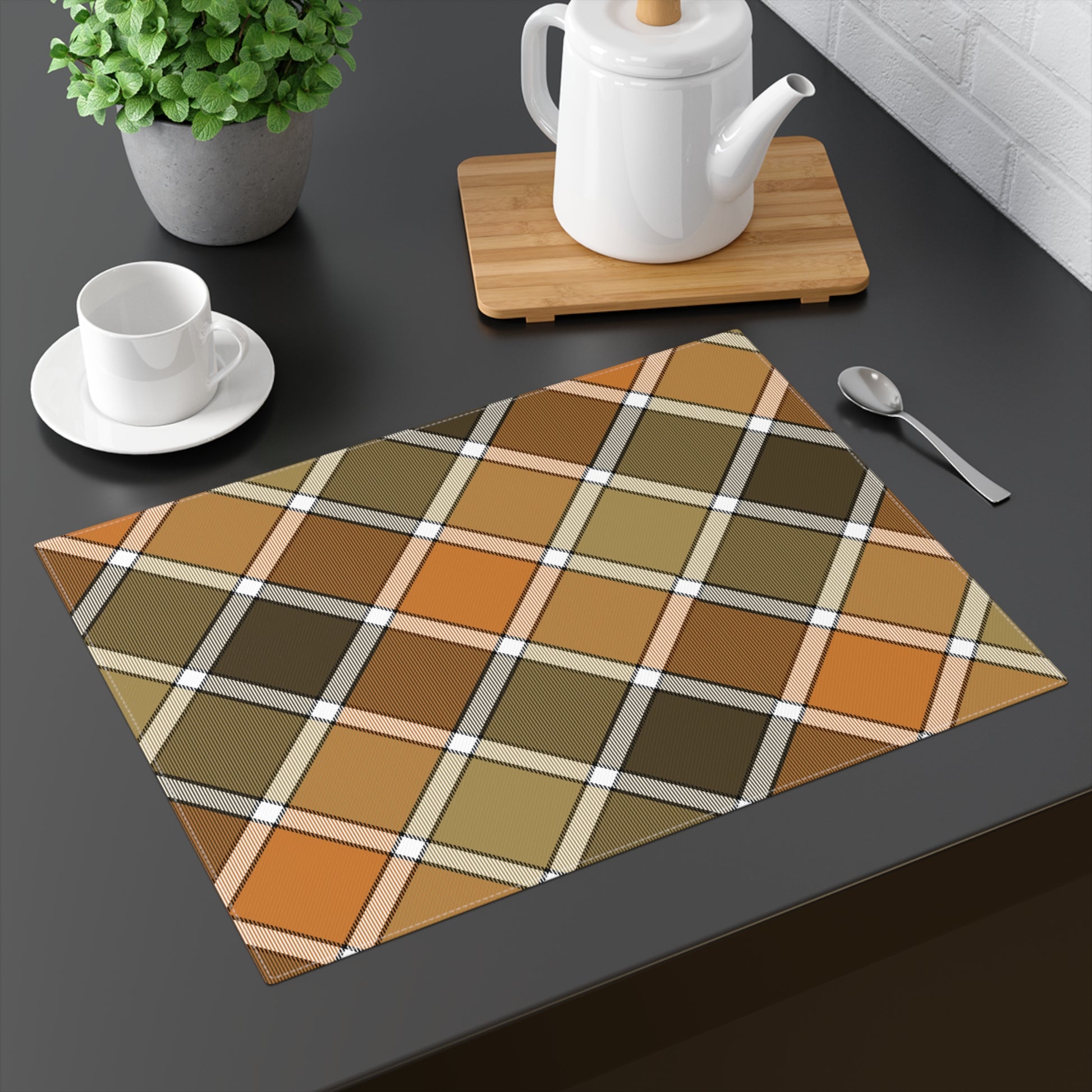 Fall Plaid placemat has plaid patterned colors with brown, orange, and green colors. The placemat sits on a dark surface with a teapot, cup, and spoon.