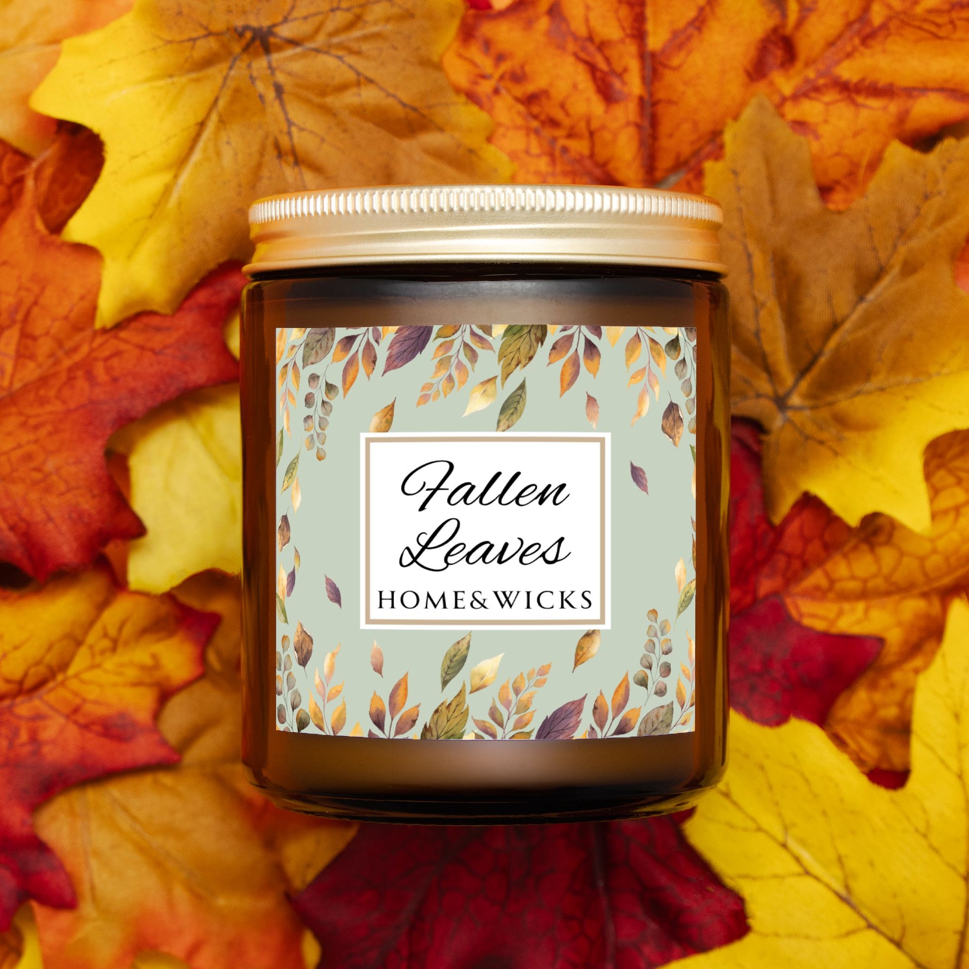 Amber glass candle jar with gold lid with a brown, gold and green 'Fallen Leaves' label on a white background by Home & Wicks on a bed of autumn leaves.