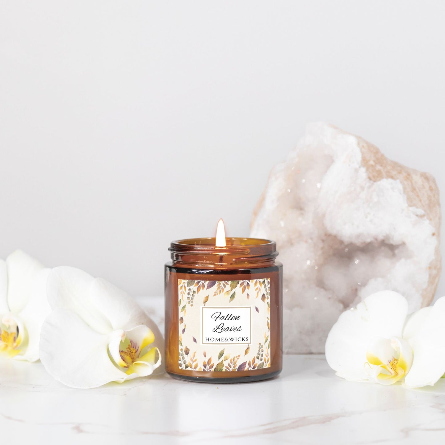 Lit candle in a glass jar with a decorative Fallen Leaves label, surrounded by white flowers and a decorative rock on a light background.