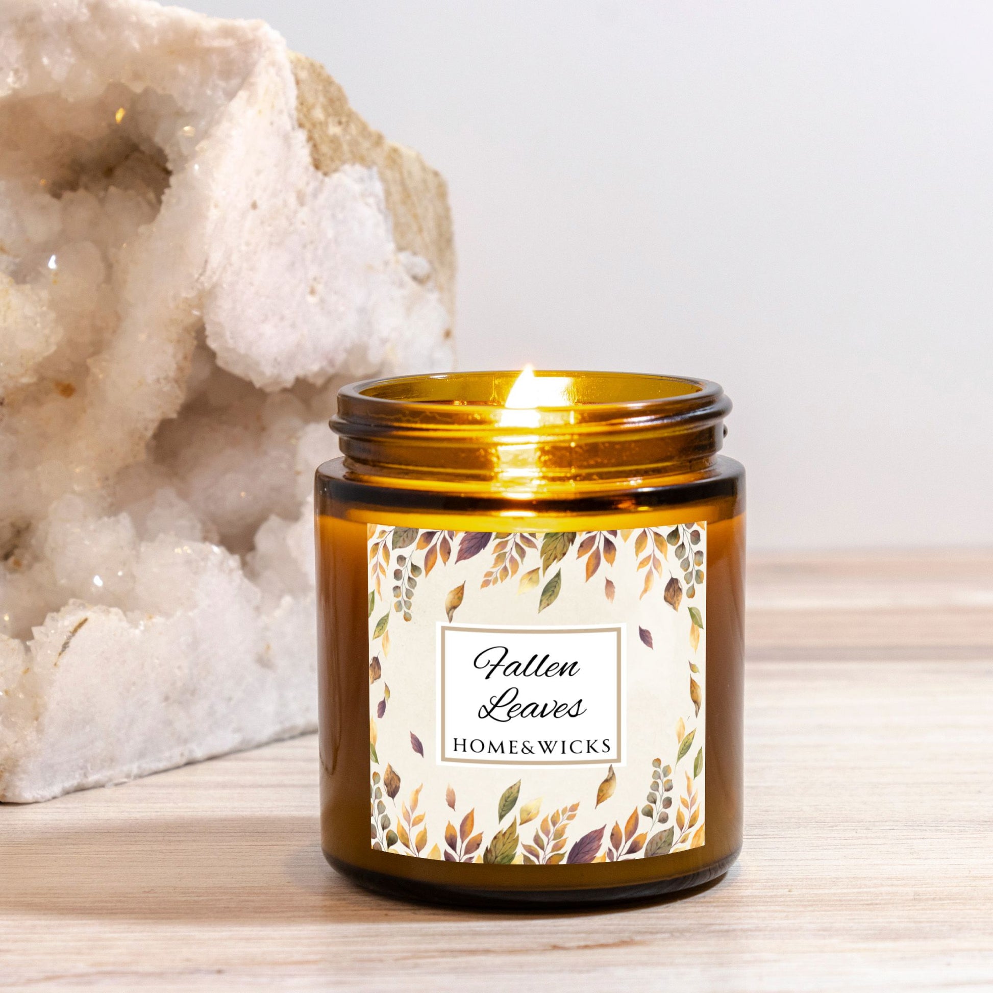 Lit candle in an amber candle jar labeled 'Fallen Leaves' by Home & Wicks on a wooden surface with a white background.