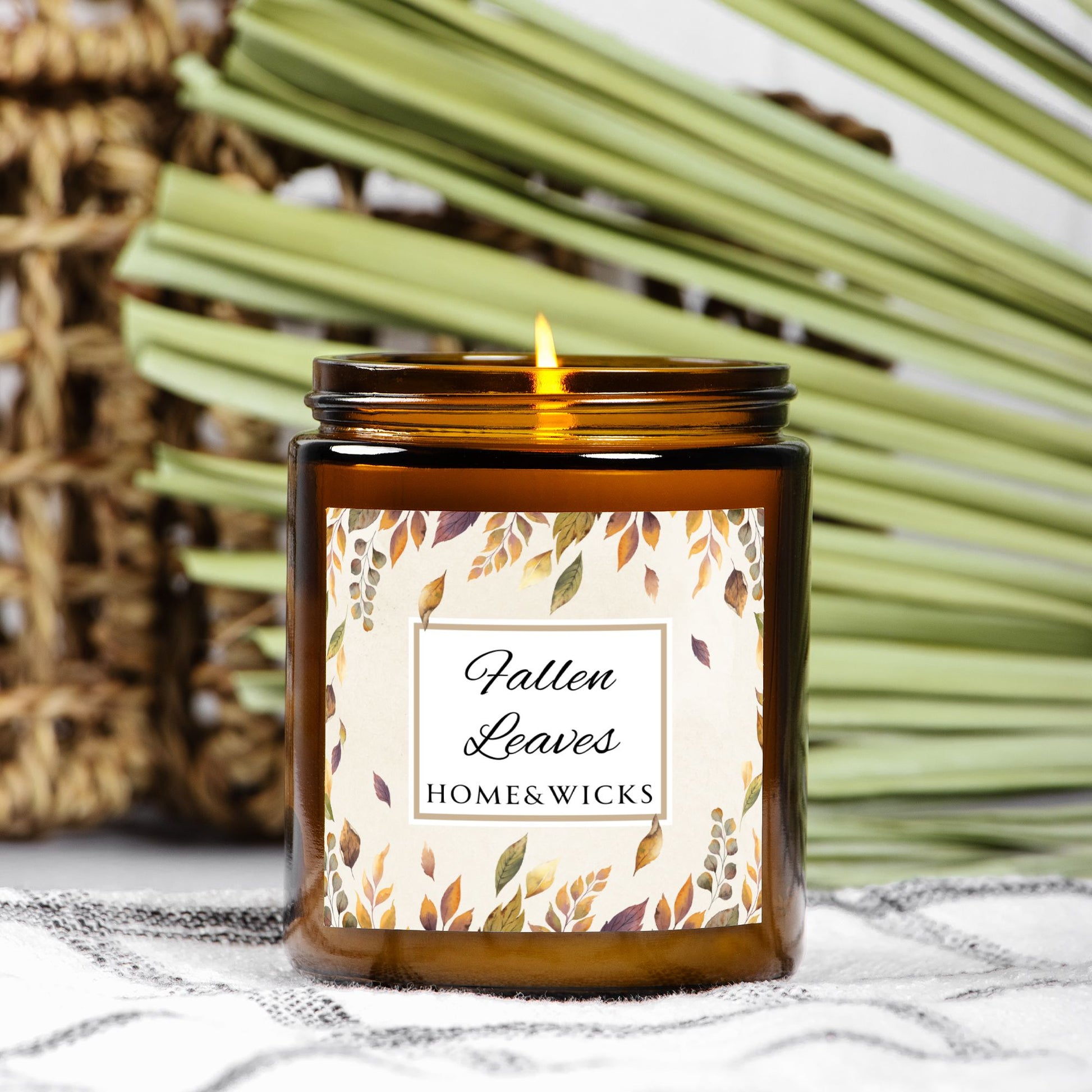 Lit candle in an amber jar with a 'Fallen Leaves' label on a textured surface with green leaves in the background.