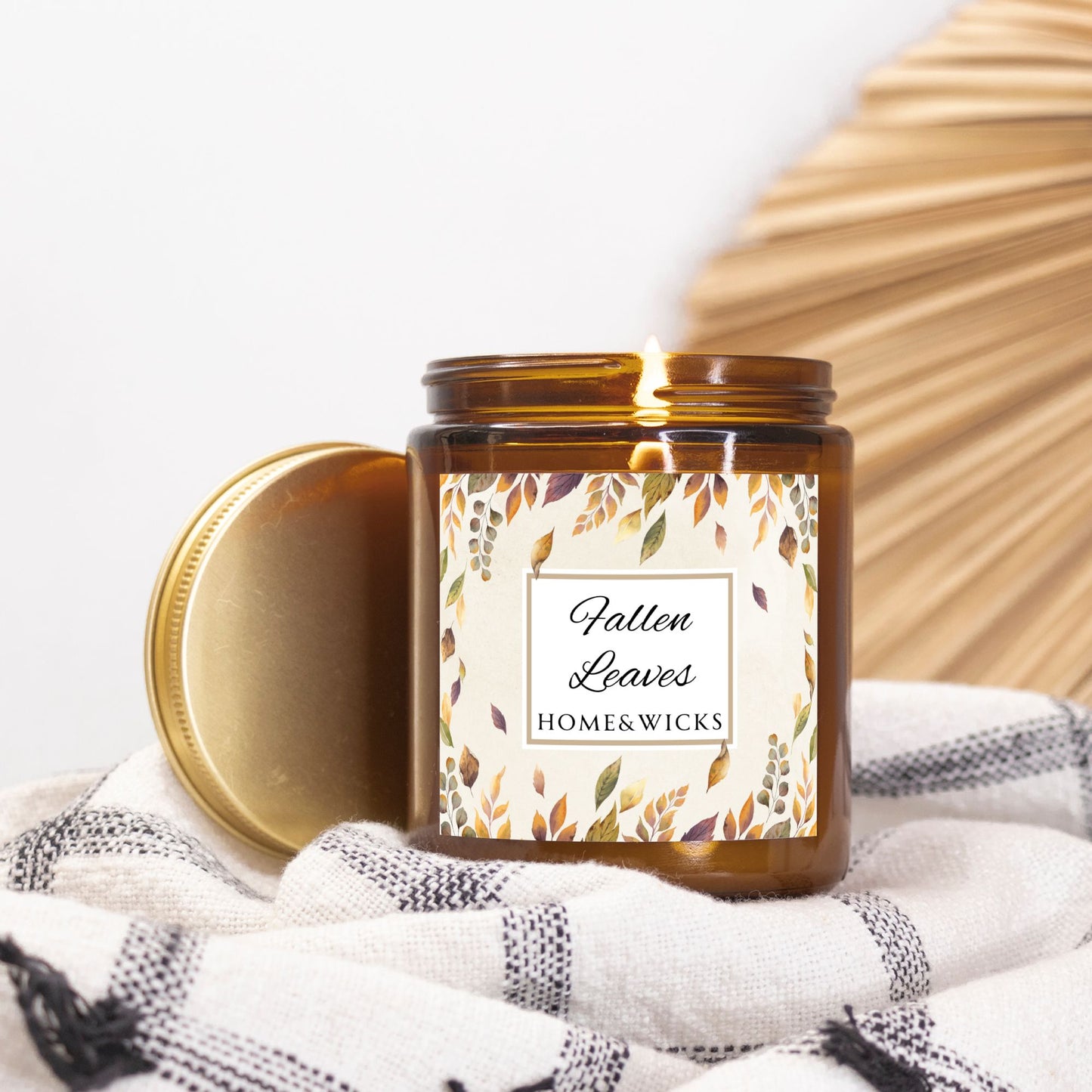 Lit candle in an amber jar with gold lid and with 'Fallen Leaves' label on a white and black checkered cloth with a wooden surface in the background.