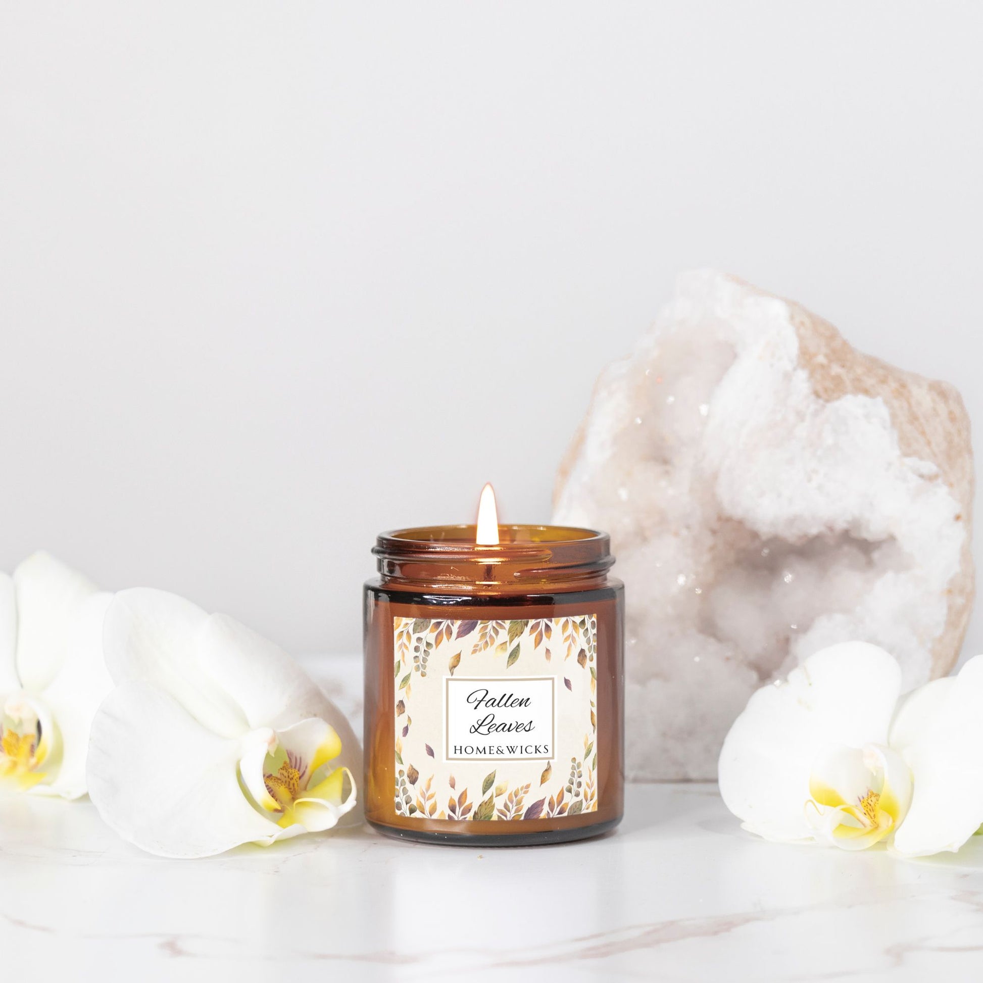 Fallen Leaves candle in an amber jar with a decorative label, surrounded by white flowers and a decorative rock on a light background.